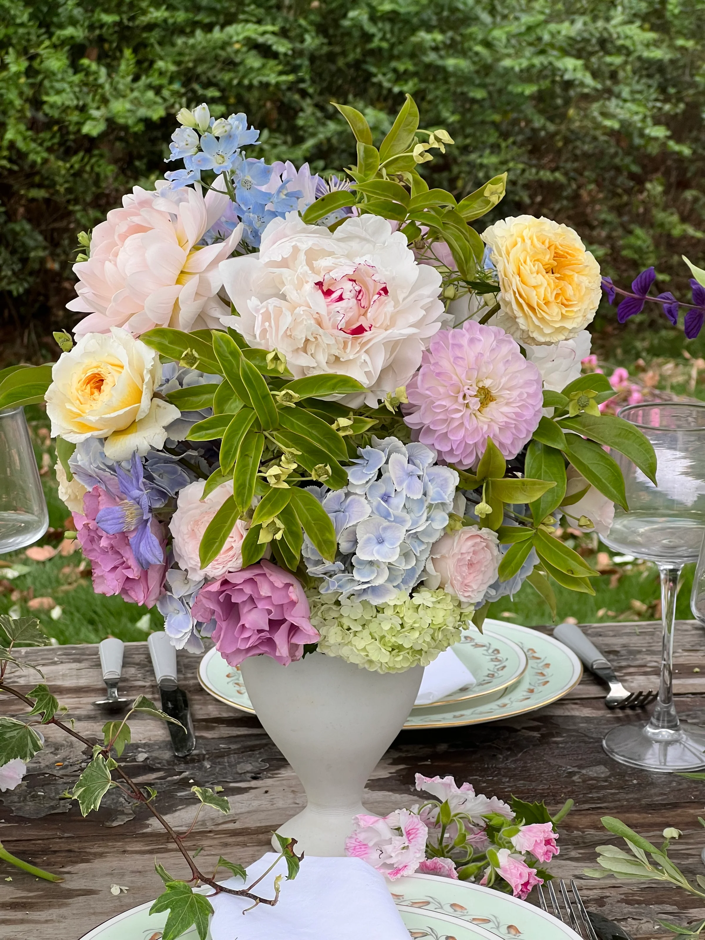 Pastel garden compote centerpiece of peonies, dahlias, and hydrangeas in a white footed vase on a rustic table