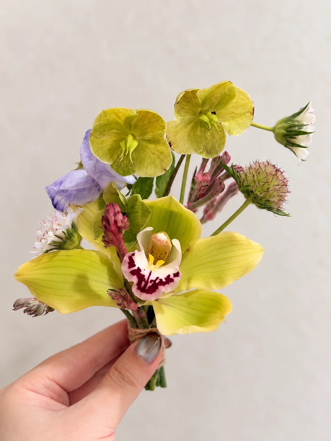 Hand holding a small floral piece with green orchids, purple flowers, and textured buds