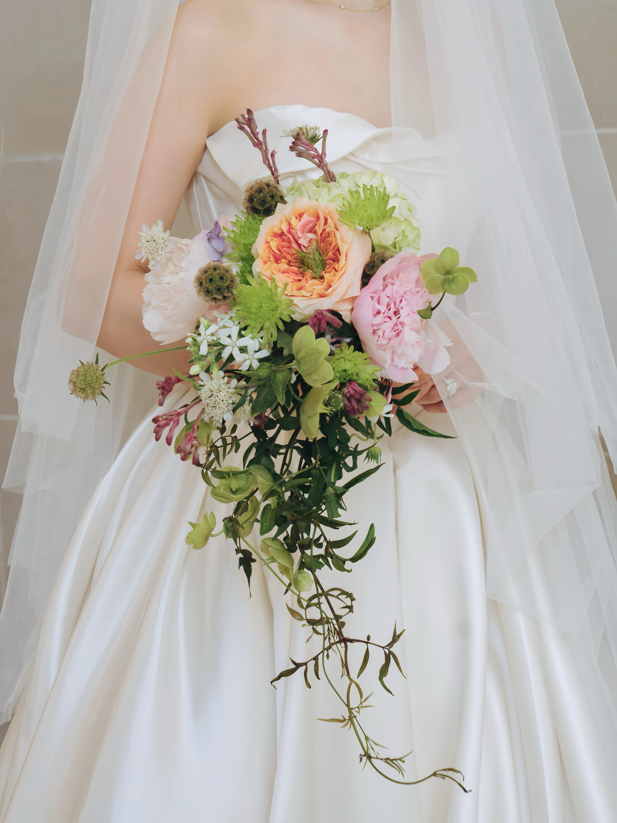 Bride holding a cascading bouquet of peach and pink flowers, green accents, and trailing foliage against an ivory gown