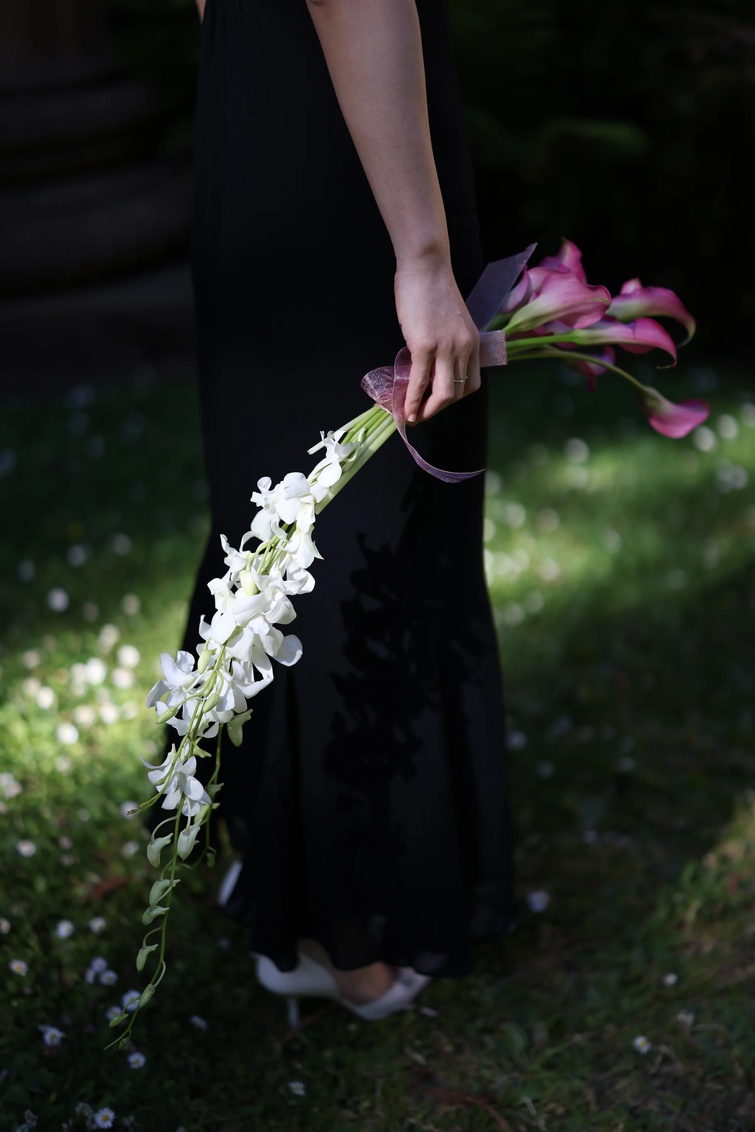 Person in a black dress holding pink calla lilies and trailing white orchids in a garden