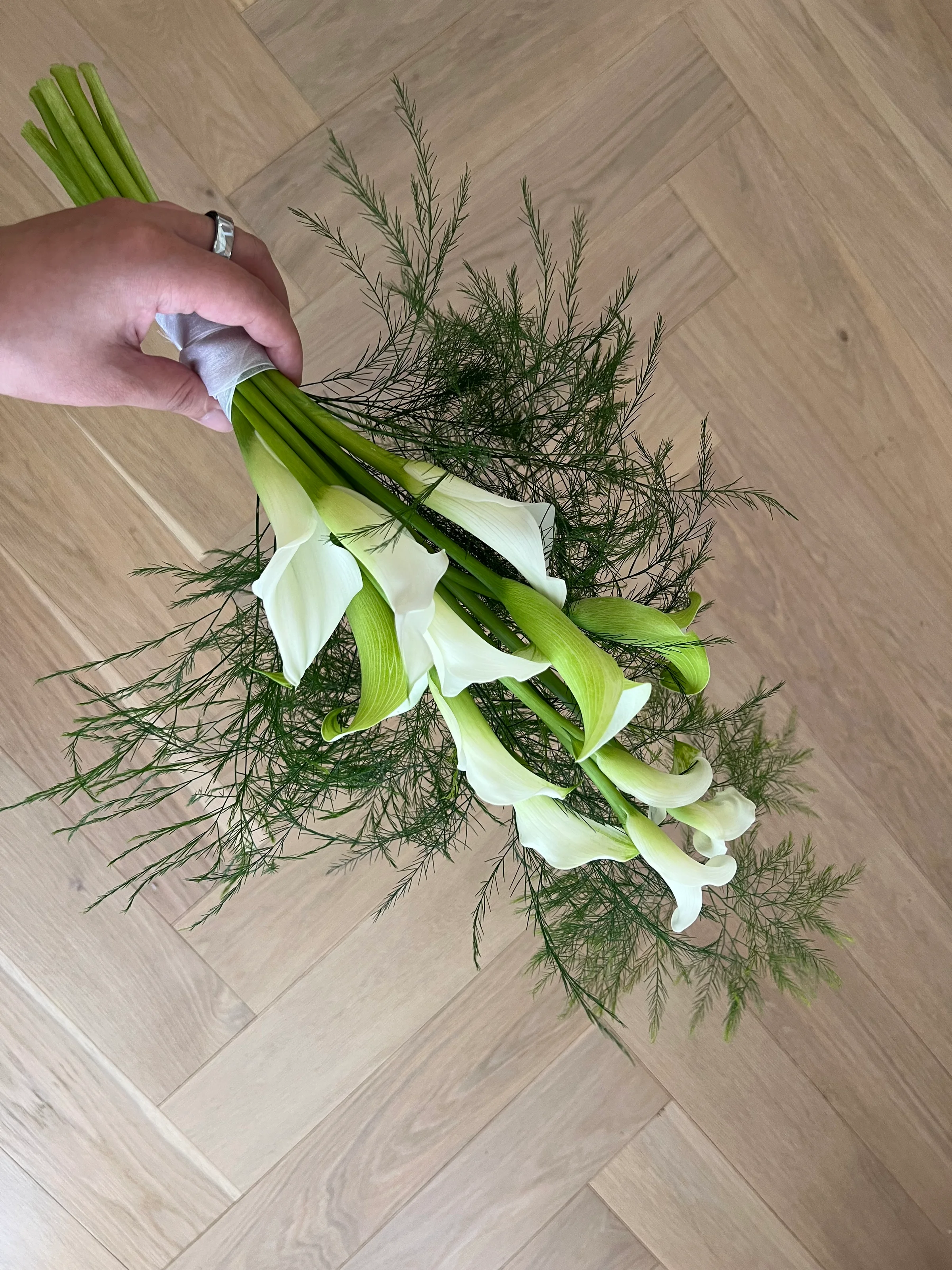 Hand holding white calla lilies and feathery greenery above a herringbone wood floor