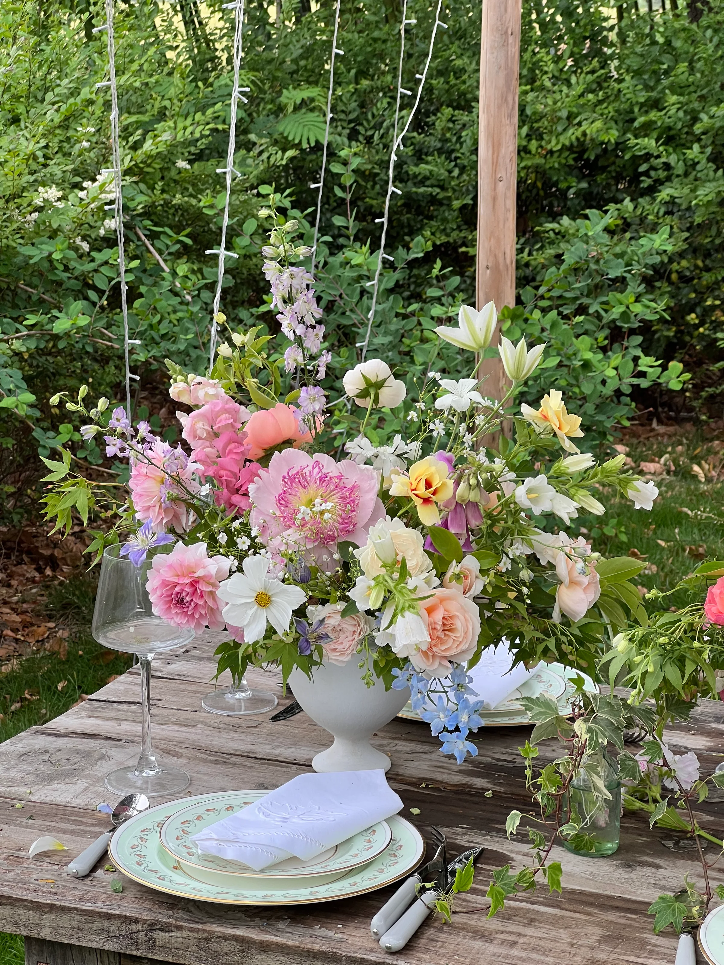 Pink, peach, and white garden centerpiece in a white footed vase beside a place setting on an outdoor wooden table