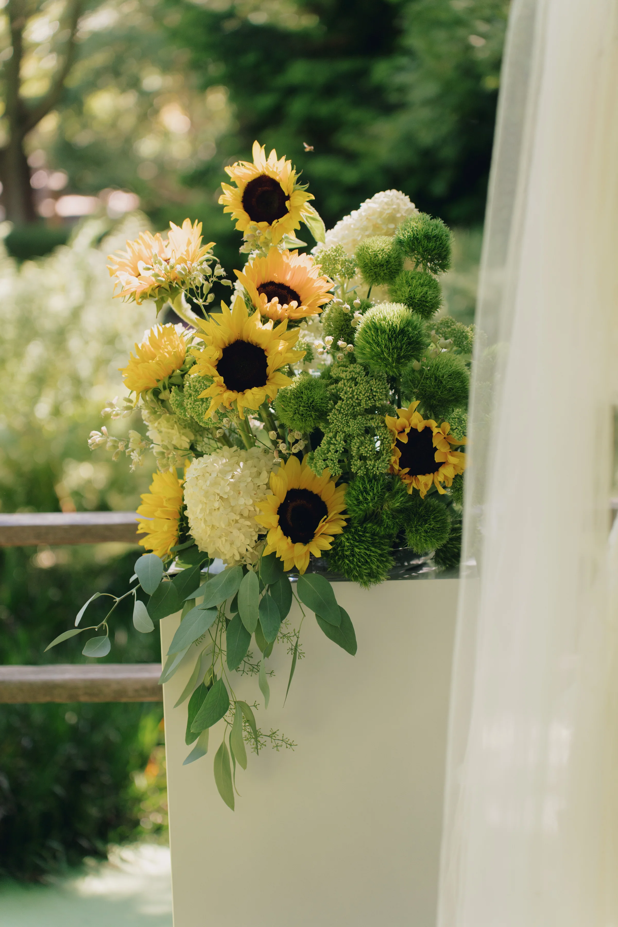 Sunflowers and pale blooms with trailing greenery arranged beside white ceremony draping