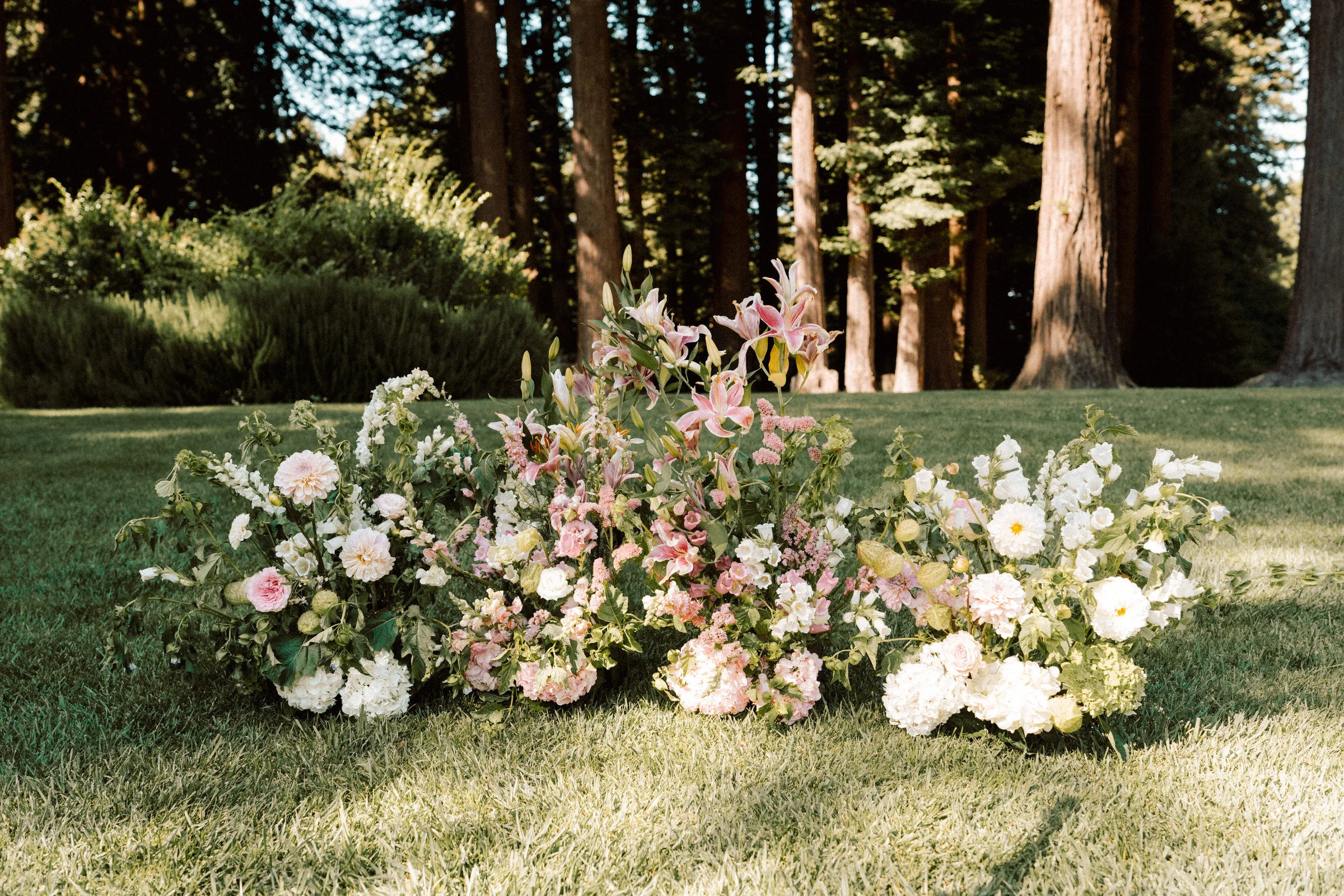 Low garden-style ceremony arrangements in white, blush, and lavender on a lawn beneath trees