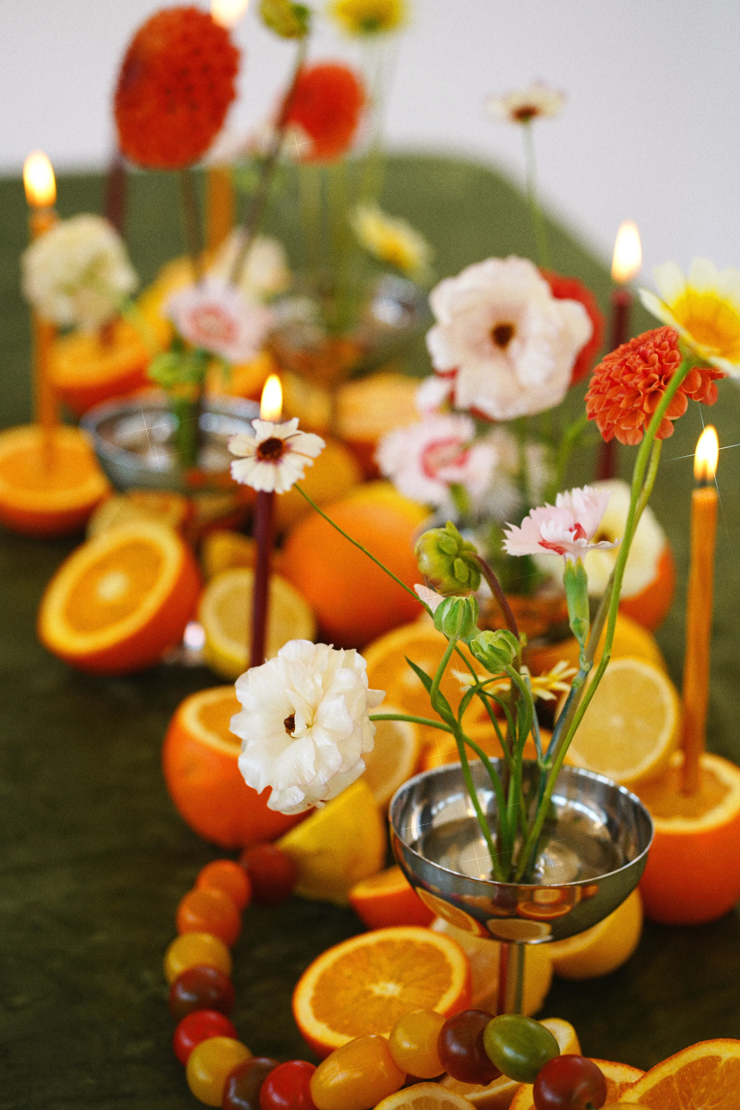 Orange dahlias and pale flowers in silver vessels among sliced citrus and lit taper candles on a moss-green tablecloth