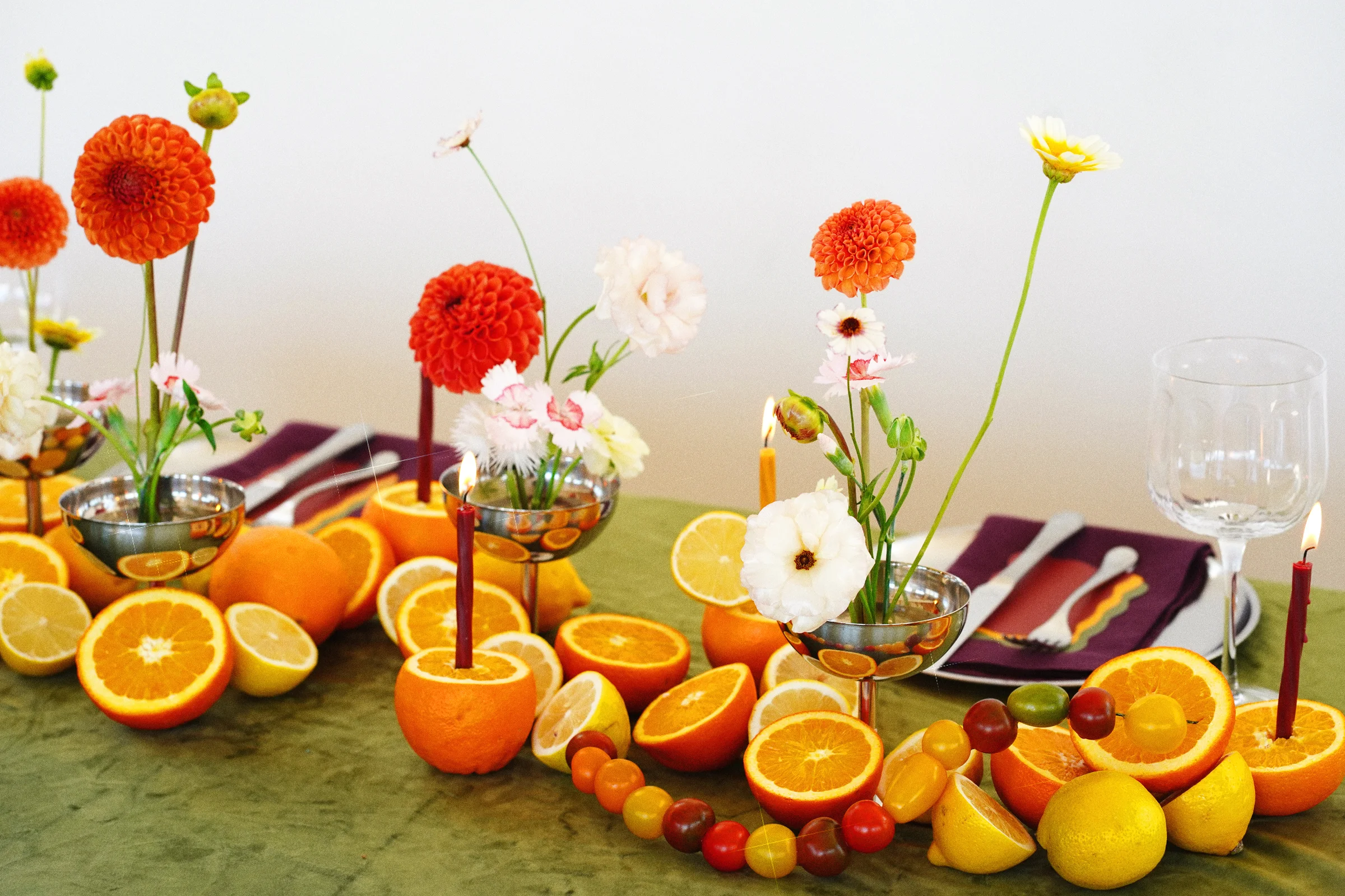 Orange dahlias, pale pink and white flowers, oranges, lemons, and cherry tomatoes beside a burgundy napkin and clear stemware