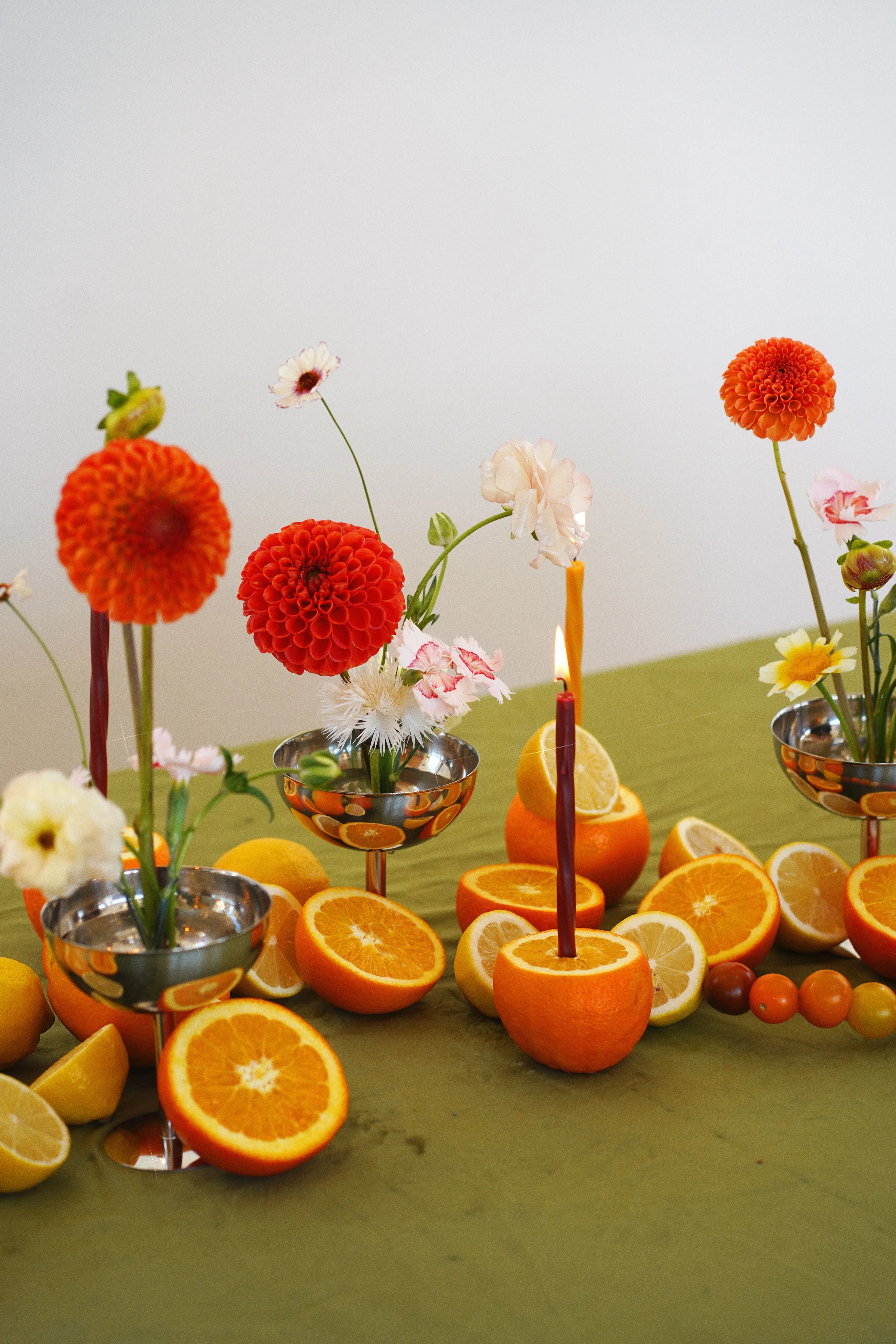 Tall orange dahlias and delicate pale blooms above halved oranges, lemons, cherry tomatoes, and a burgundy taper candle