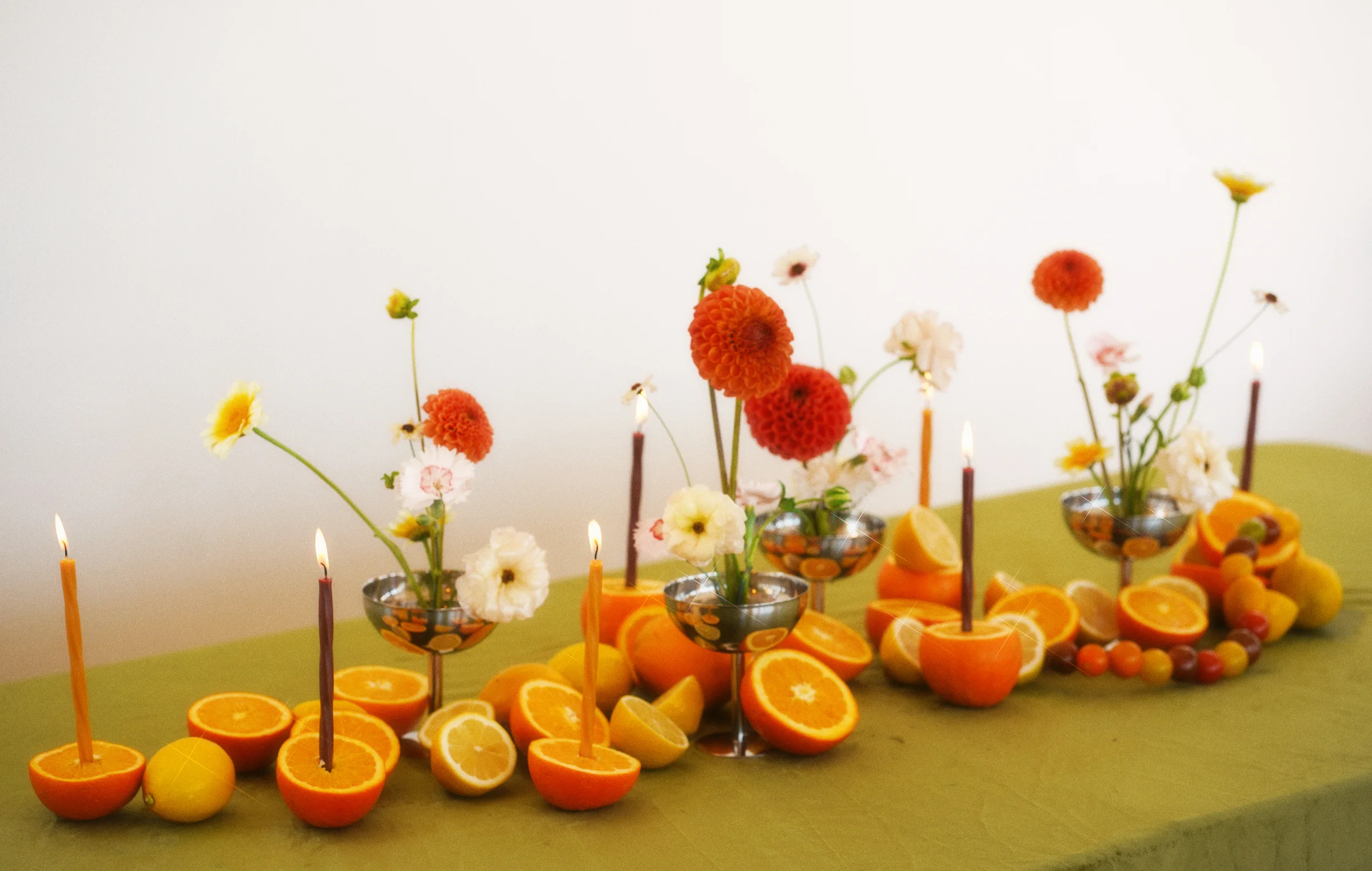 Wide view of floral arrangements and citrus fruit across a green-covered table, with taper candles set into orange halves