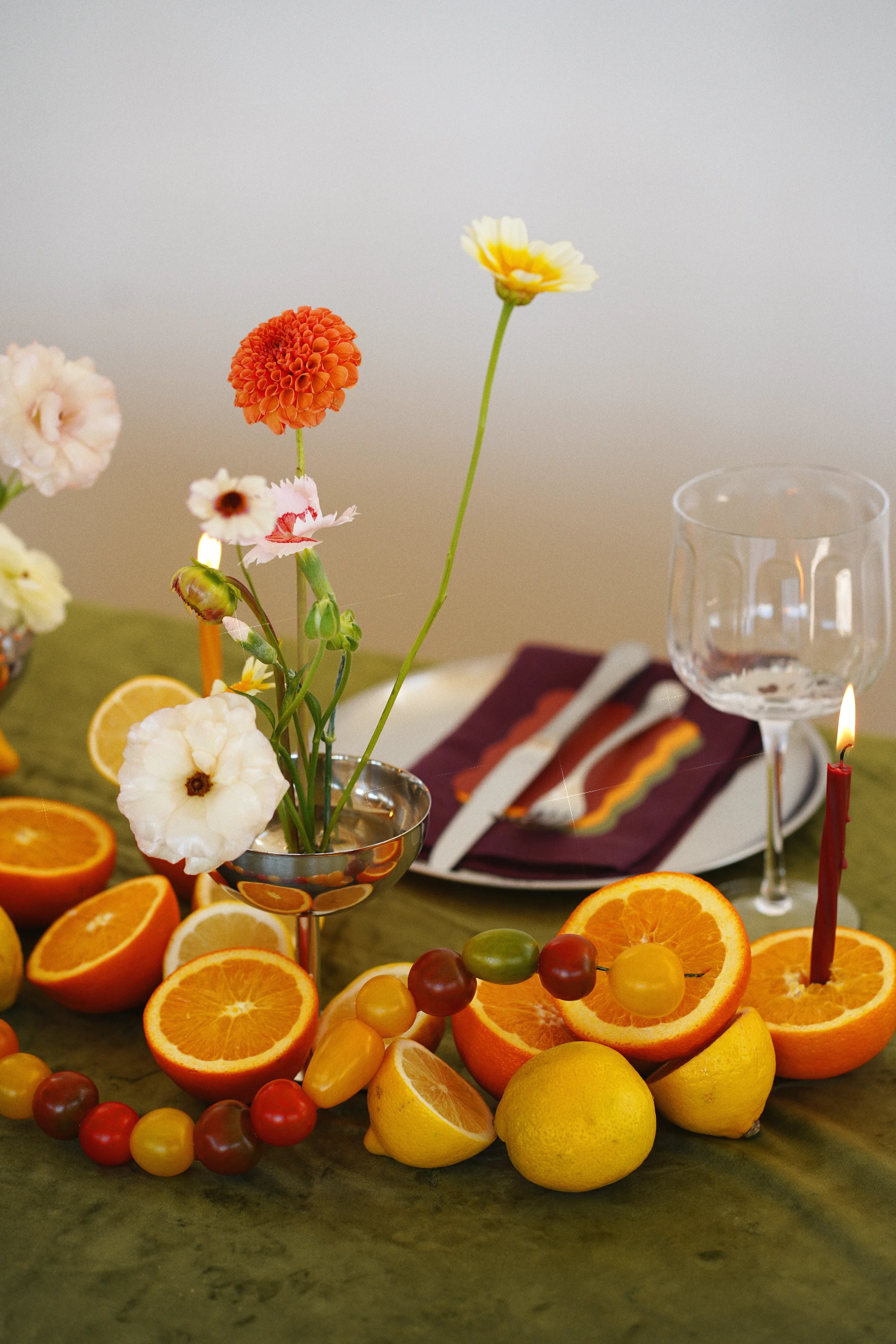 White and blush flowers in a silver vessel beside oranges, lemons, cherry tomatoes, and a white plate with a burgundy napkin