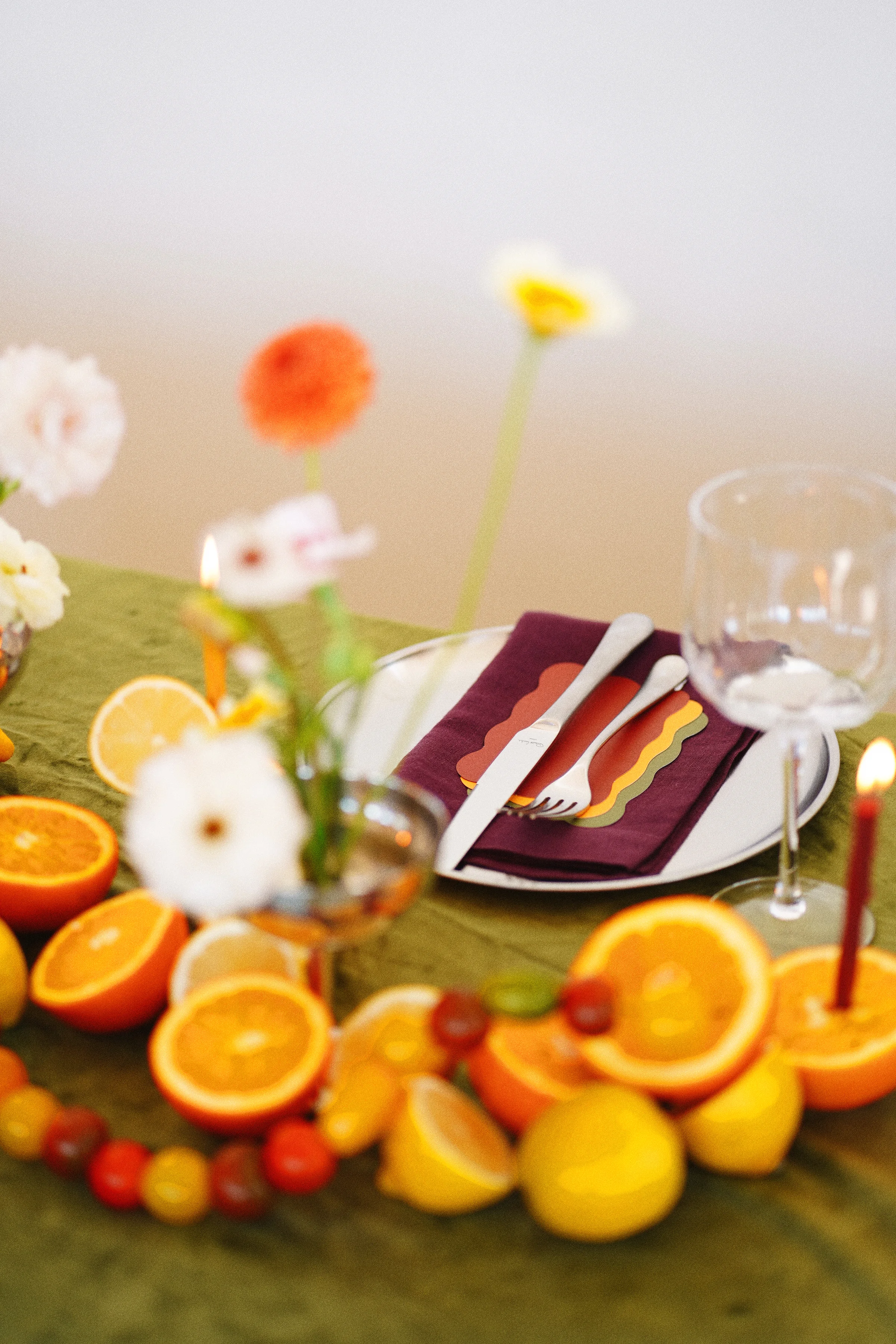 Soft-focus flowers and citrus frame a place setting with a burgundy napkin, colorful scalloped accents, and clear stemware