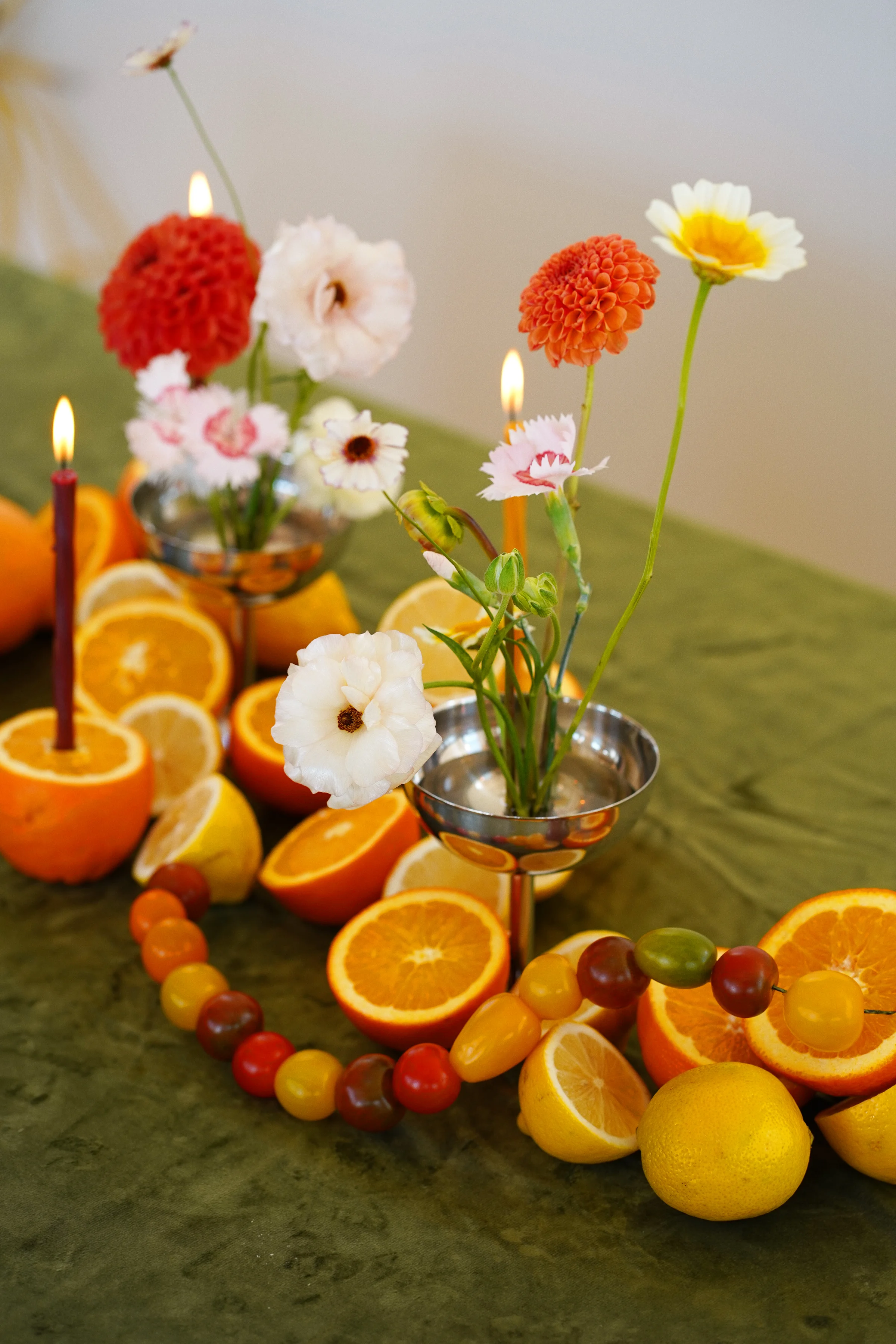 Orange dahlias and airy pale flowers rise above silver vessels surrounded by citrus halves and a curved row of cherry tomatoes