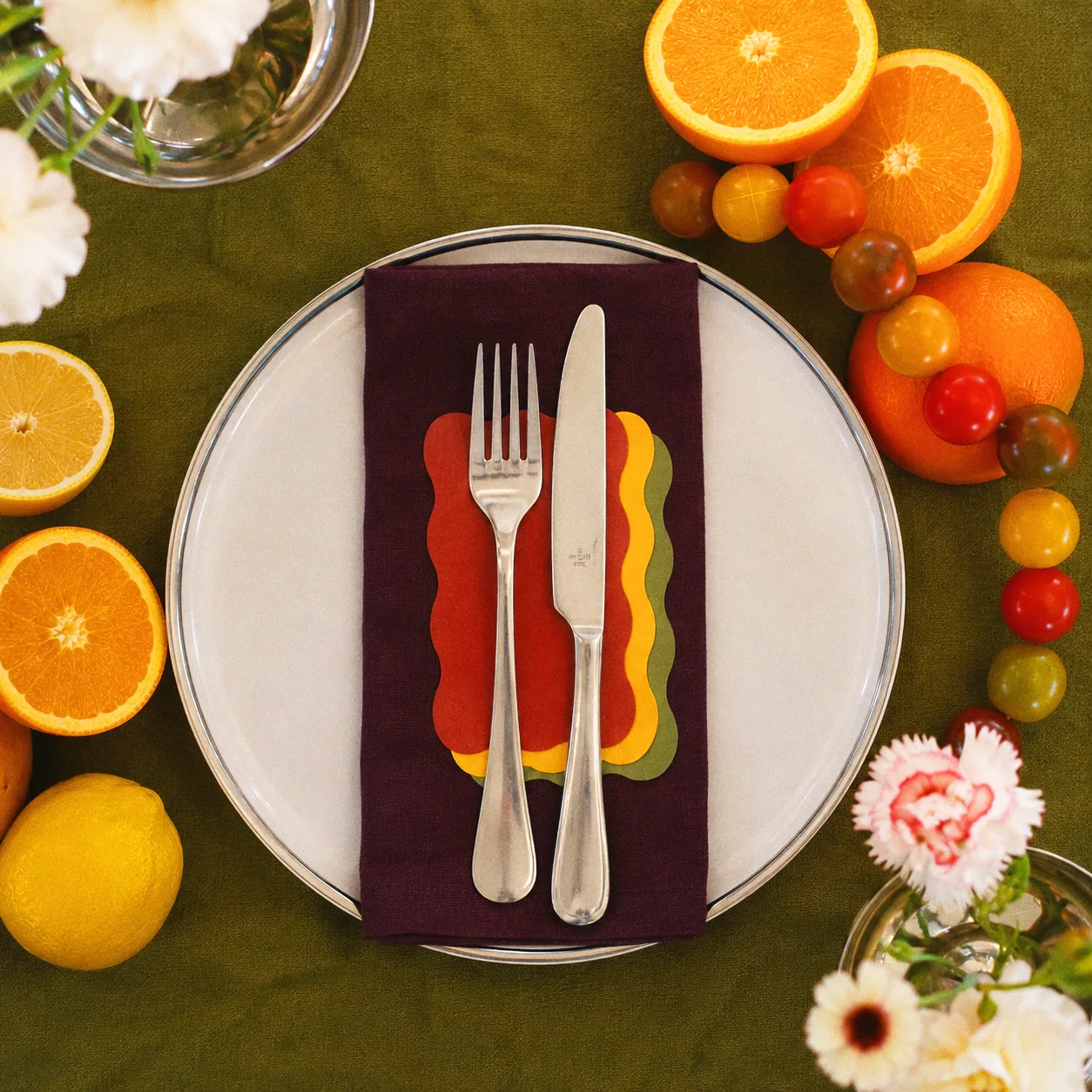 Overhead view of a white plate, burgundy napkin, silver flatware, and colorful scalloped accents framed by citrus and flowers