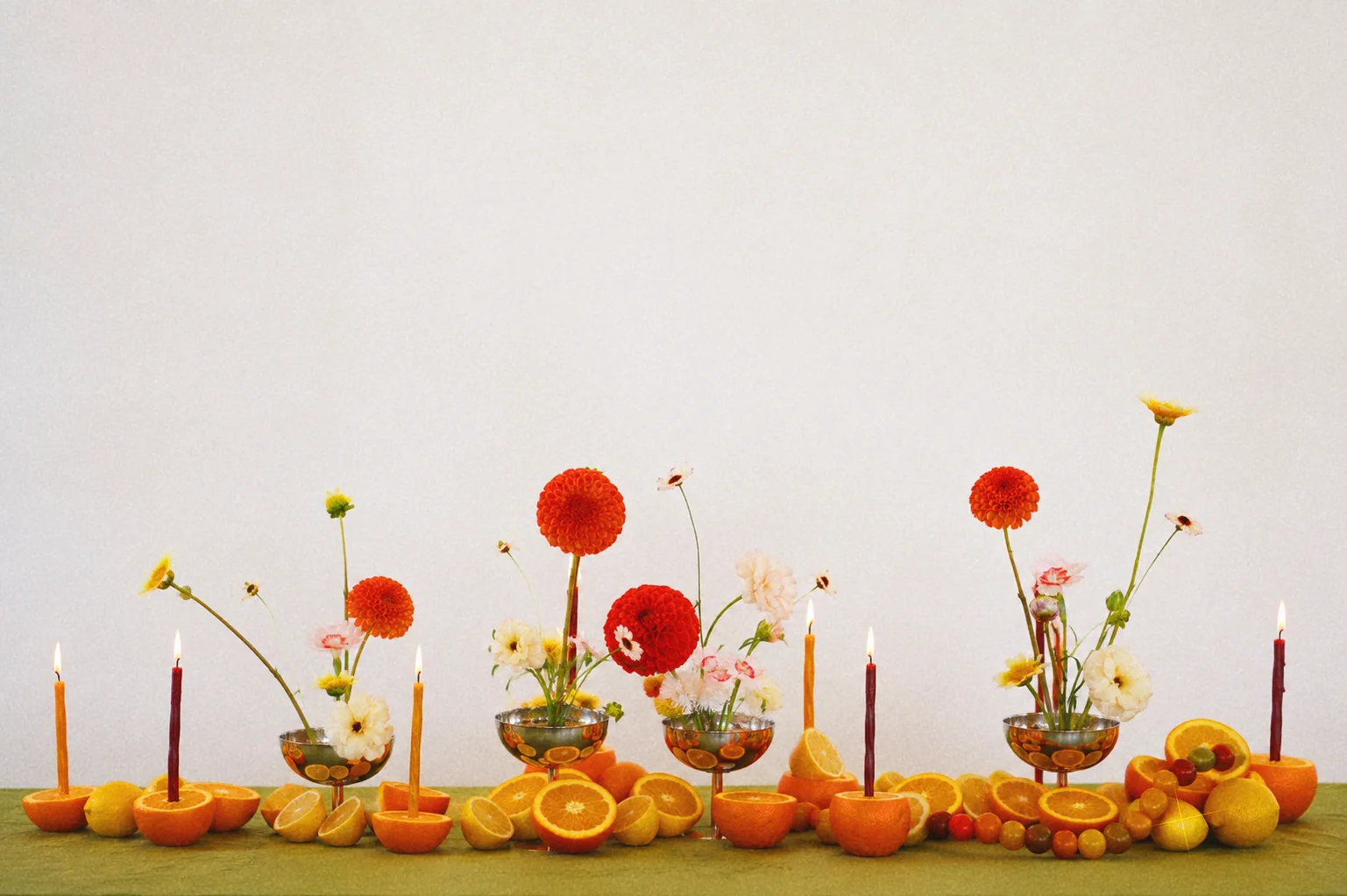Full tablescape with spaced floral arrangements, orange dahlias, pale flowers, citrus fruit, and slender candles against a white backdrop