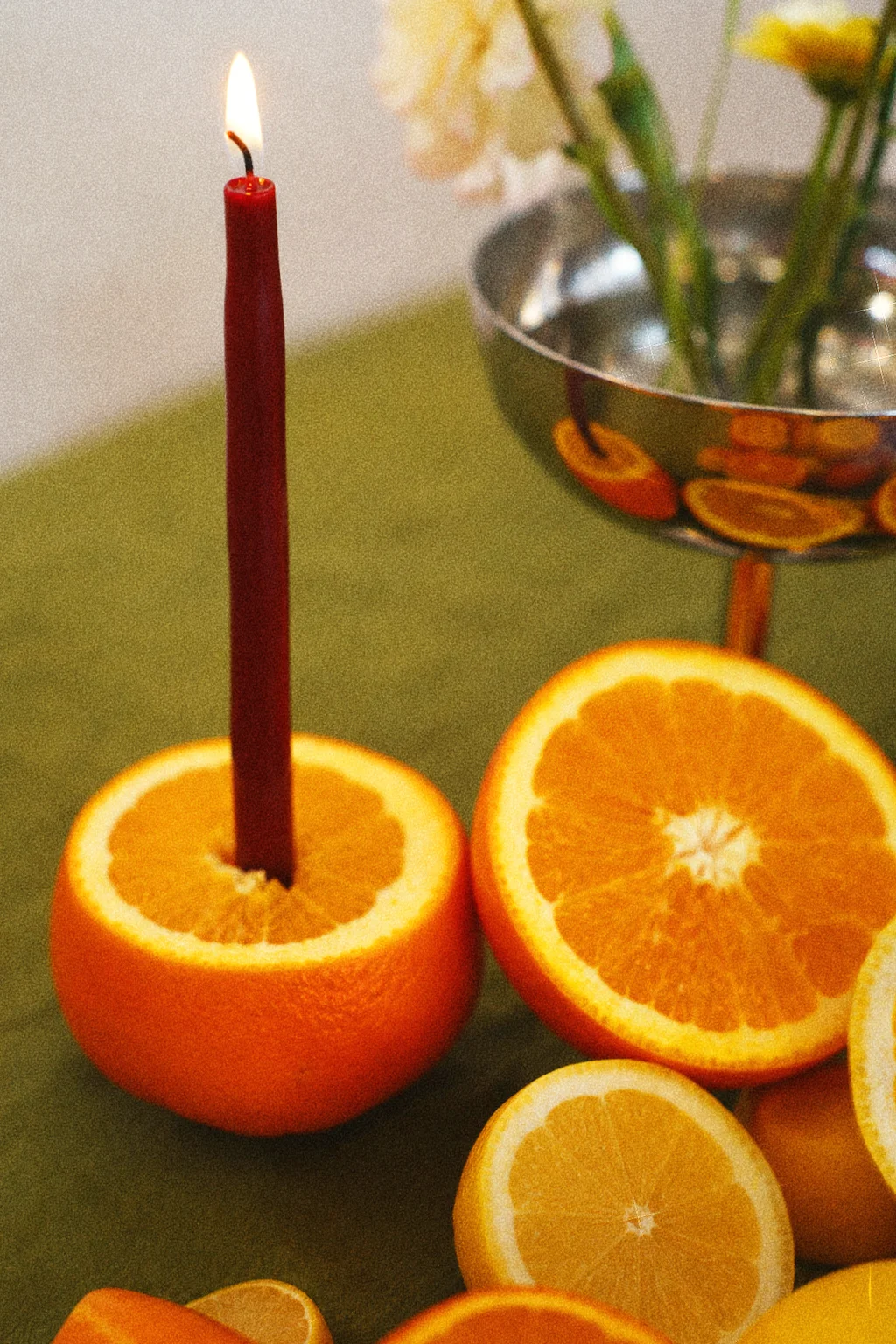 A lit burgundy taper candle set into an orange half beside sliced oranges, a lemon, and a silver floral vessel