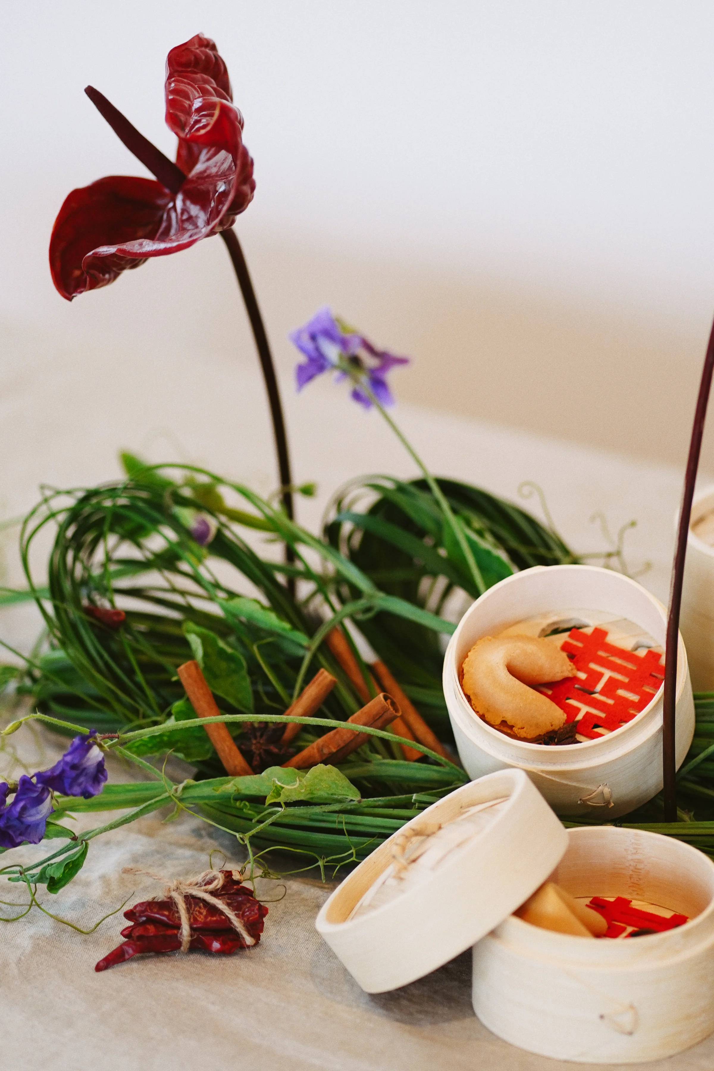 Burgundy anthuriums and purple flowers rise from looping greenery beside bamboo steamers filled with fortune cookies and red paper decorations