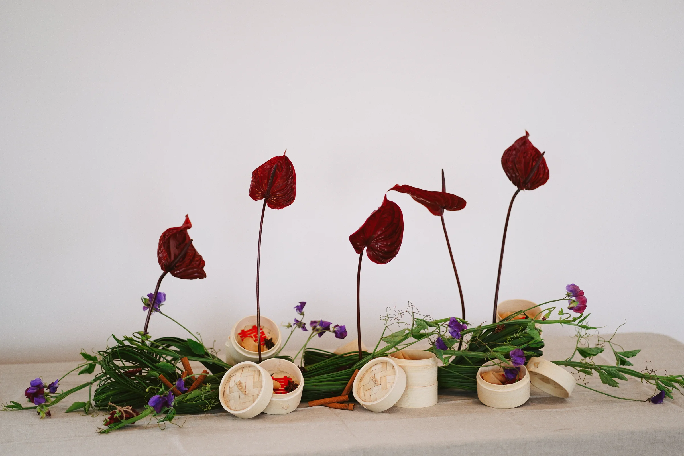 Wide floral composition with tall burgundy anthuriums, woven green leaves, purple blooms, and small bamboo steamers on a linen-covered table
