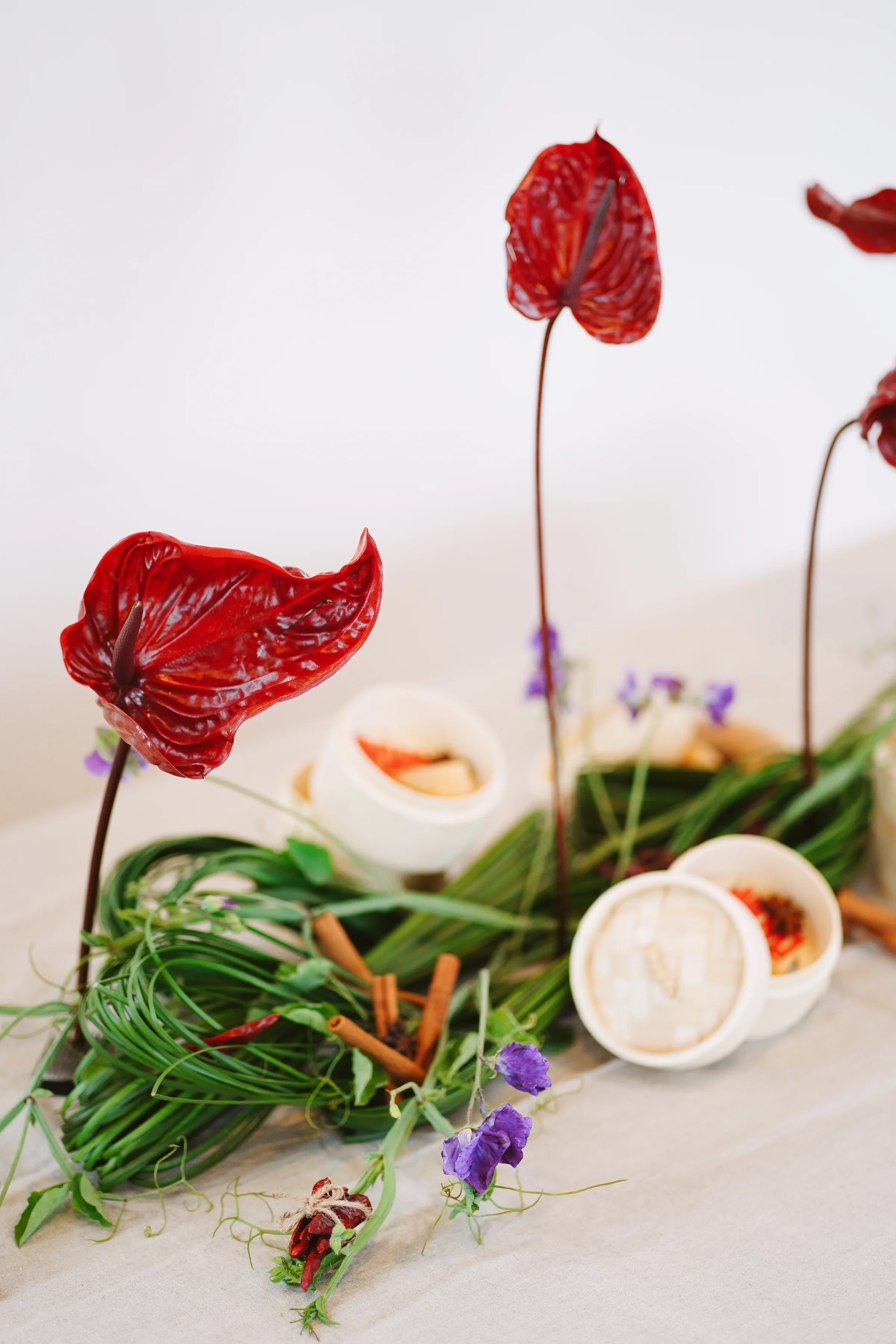 Close detail of glossy burgundy anthuriums above curled greenery, purple flowers, and open bamboo steamers