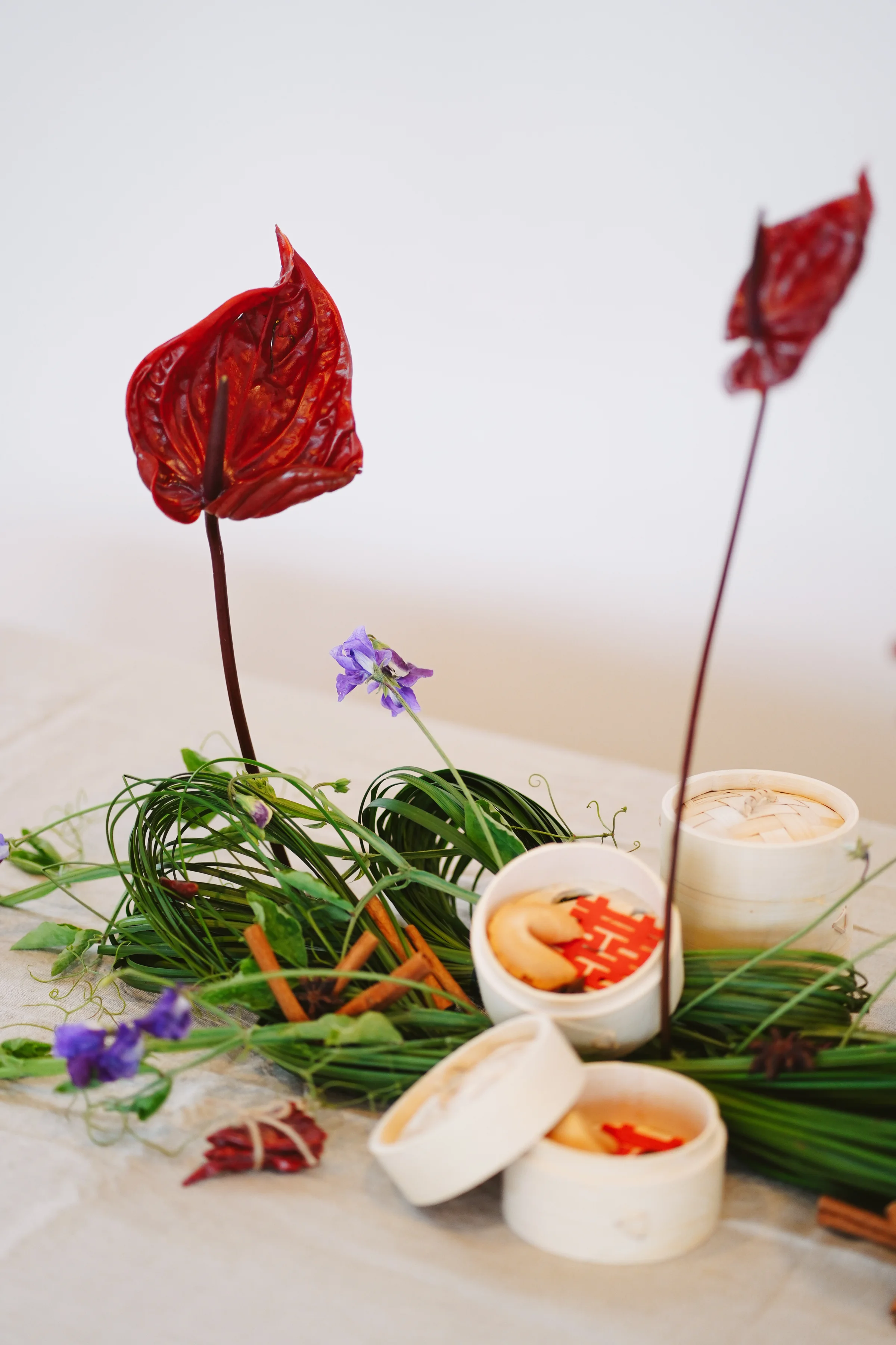 Burgundy anthuriums extend over woven greenery and purple flowers beside bamboo steamers with fortune cookies and red accents