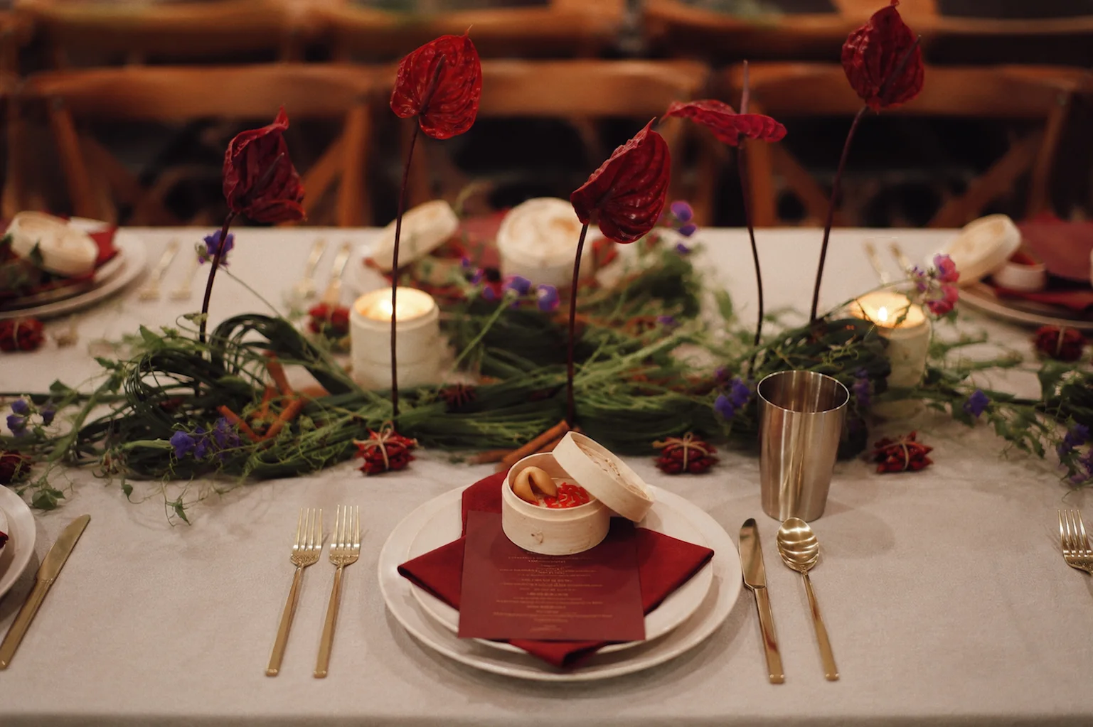 Dinner place setting with a red napkin, white plates, gold-toned flatware, and a small bamboo steamer in front of a candlelit anthurium centerpiece