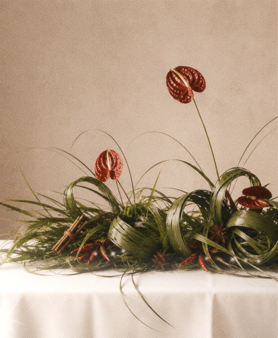 Airy burgundy anthuriums and fine grasses rise above broad looping leaves on a white-draped table against a warm neutral backdrop