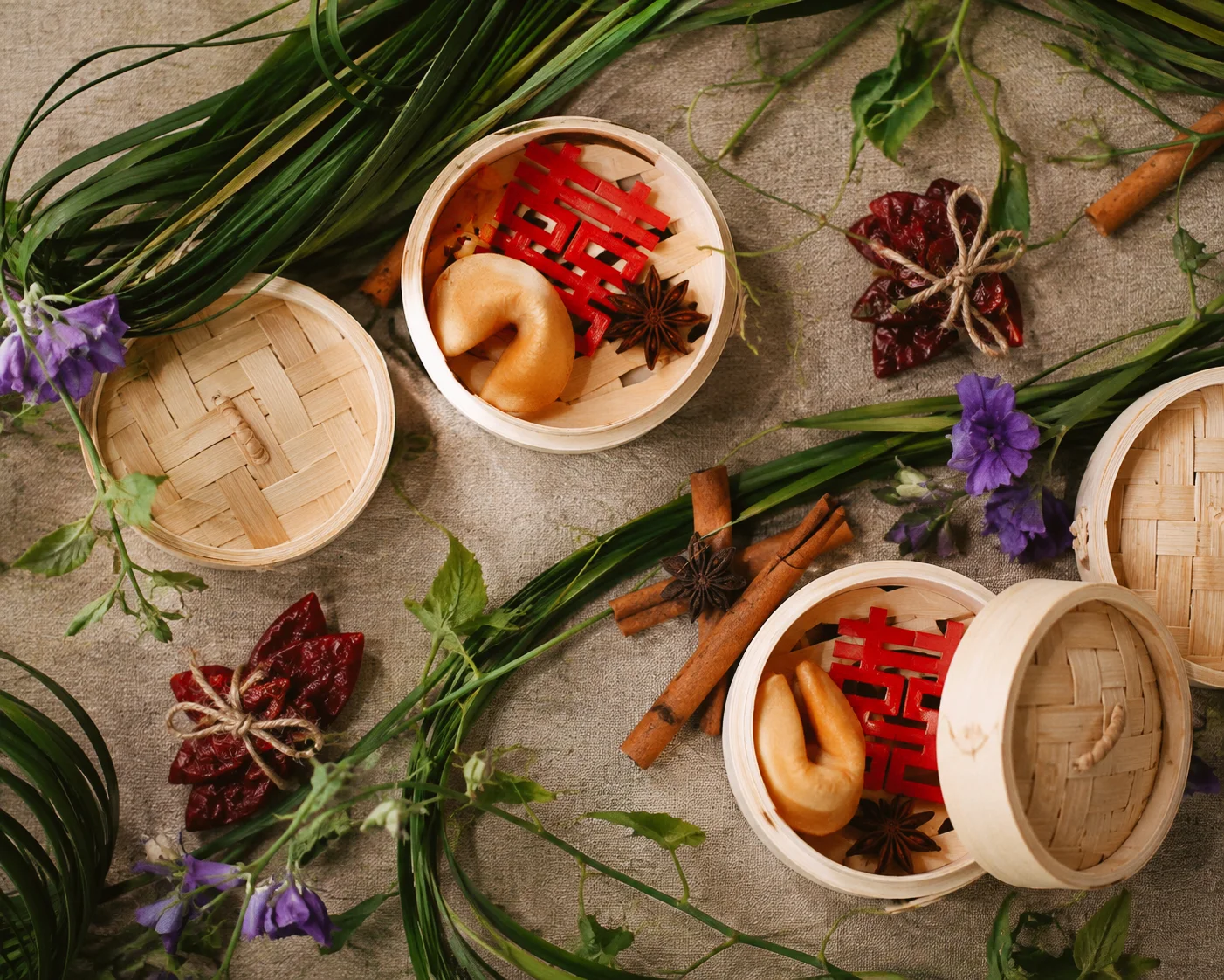 Overhead view of bamboo steamers with fortune cookies and red paper decorations among purple flowers, curled greenery, and cinnamon sticks