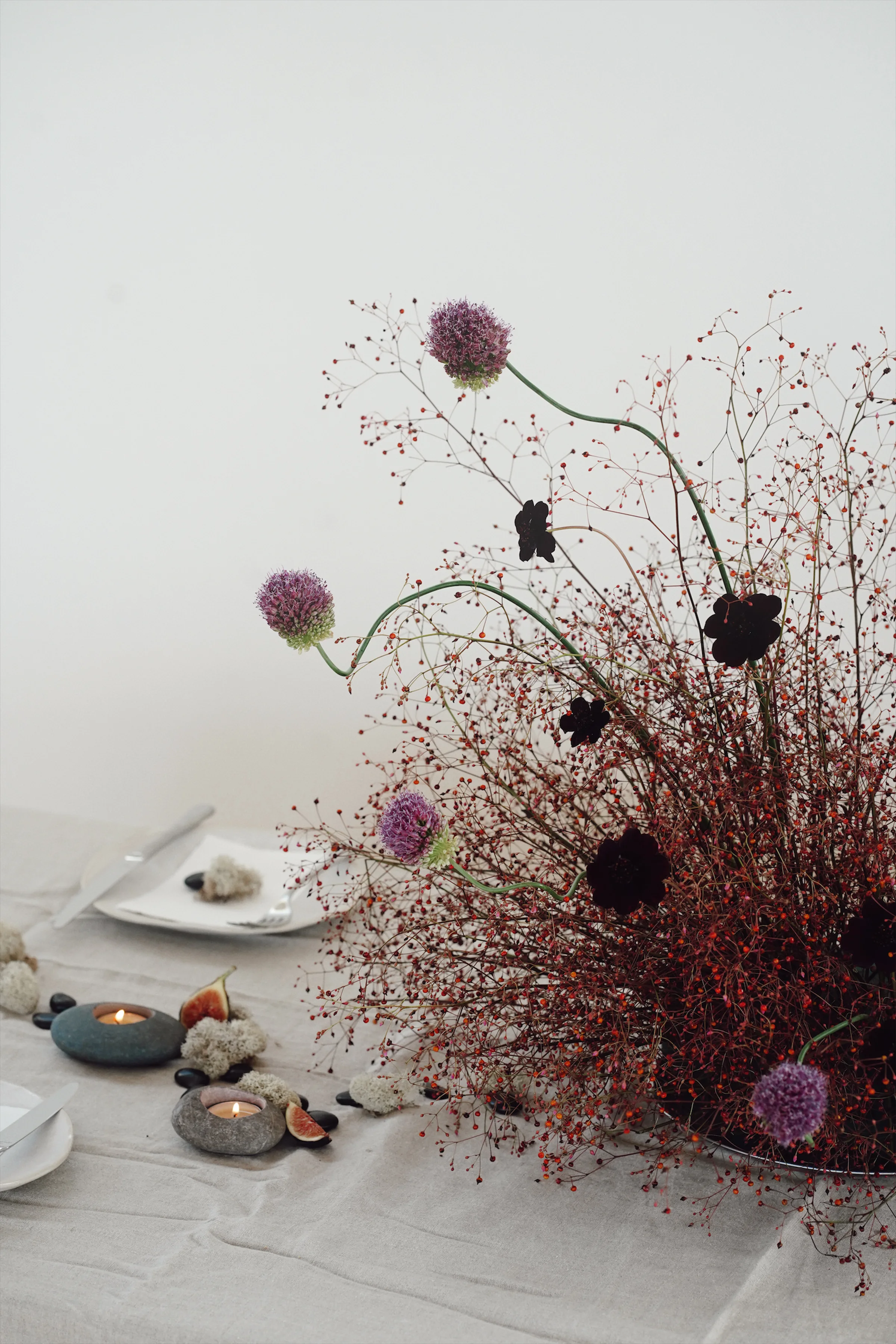 Editorial tablescape with airy red berry stems, purple alliums, dark blooms, stone candles, figs, and white place settings