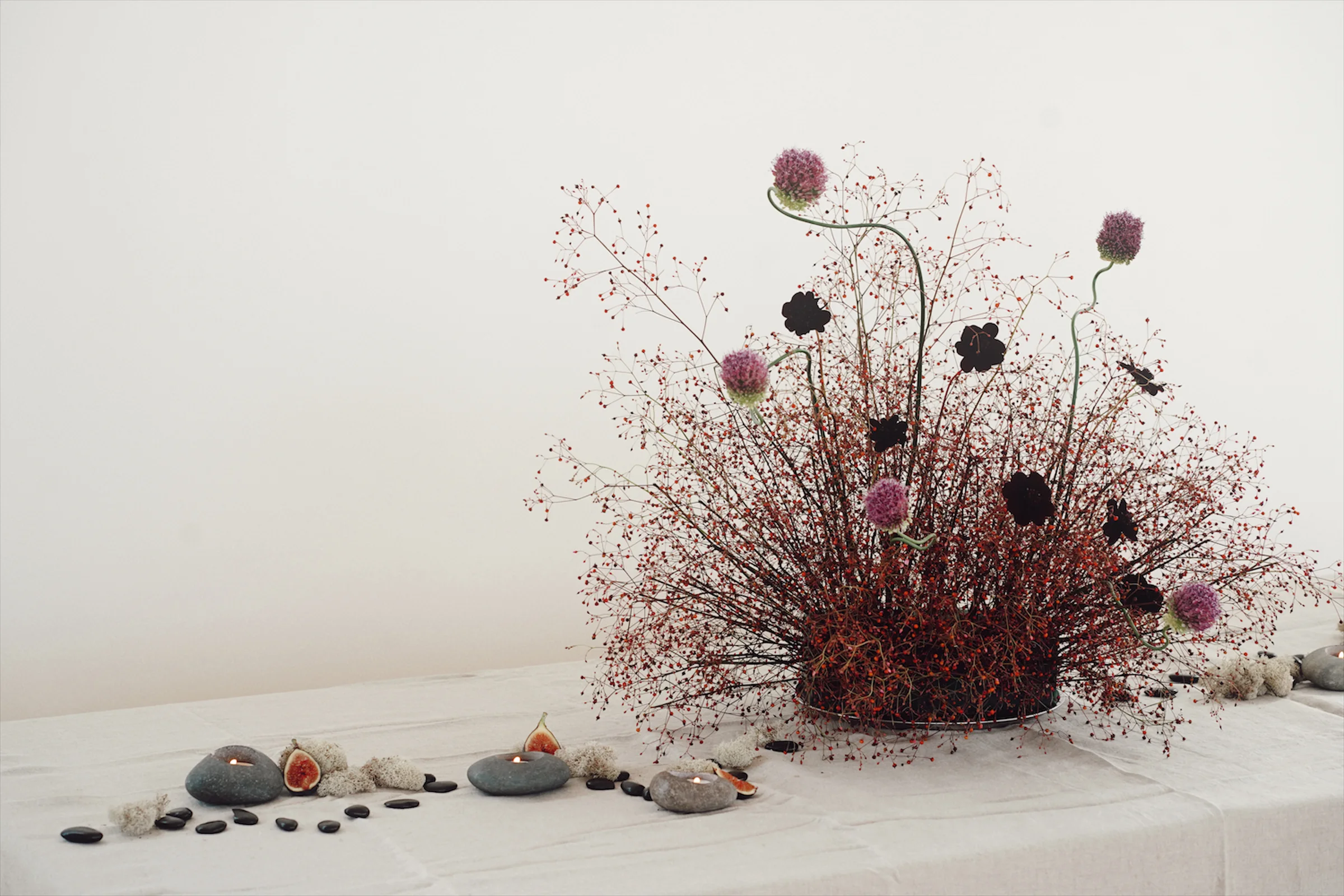 Asymmetrical floral editorial arrangement with red berry stems, purple alliums, dark blooms, figs, stone candles, and open white space