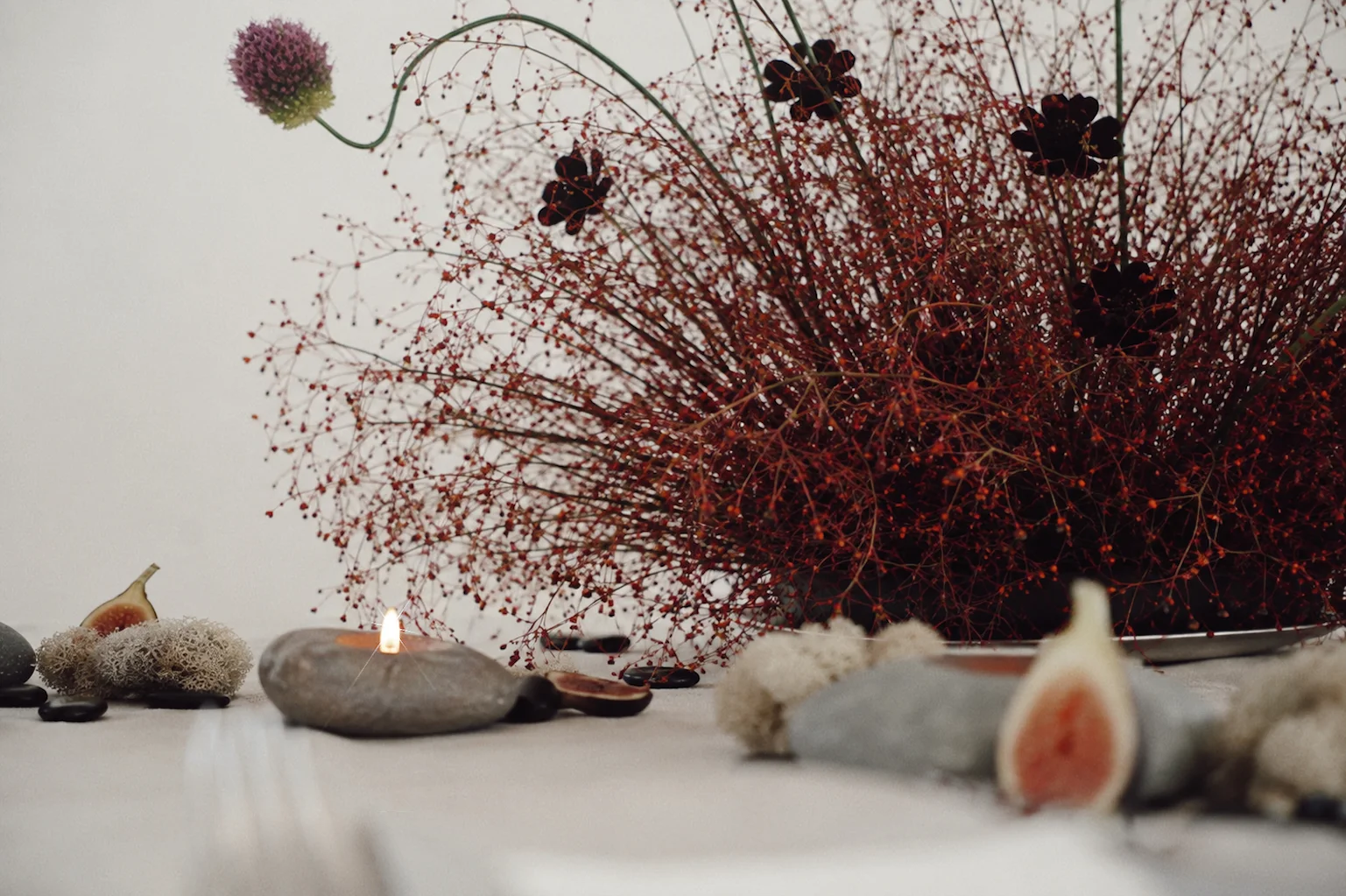 Low angle tablescape detail with a stone candle, figs, preserved moss, and the base of an airy red berry floral arrangement