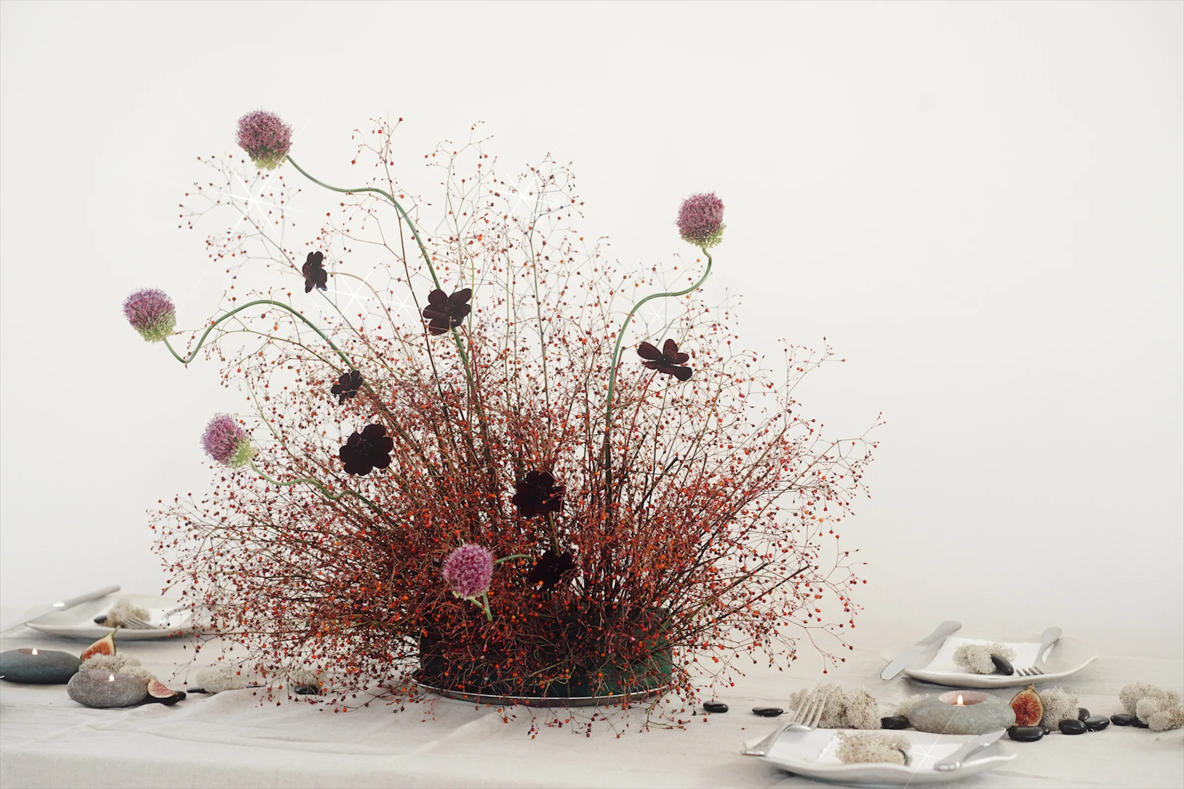 Sculptural editorial arrangement of red berry stems, purple alliums, and dark blooms framed by minimalist white place settings
