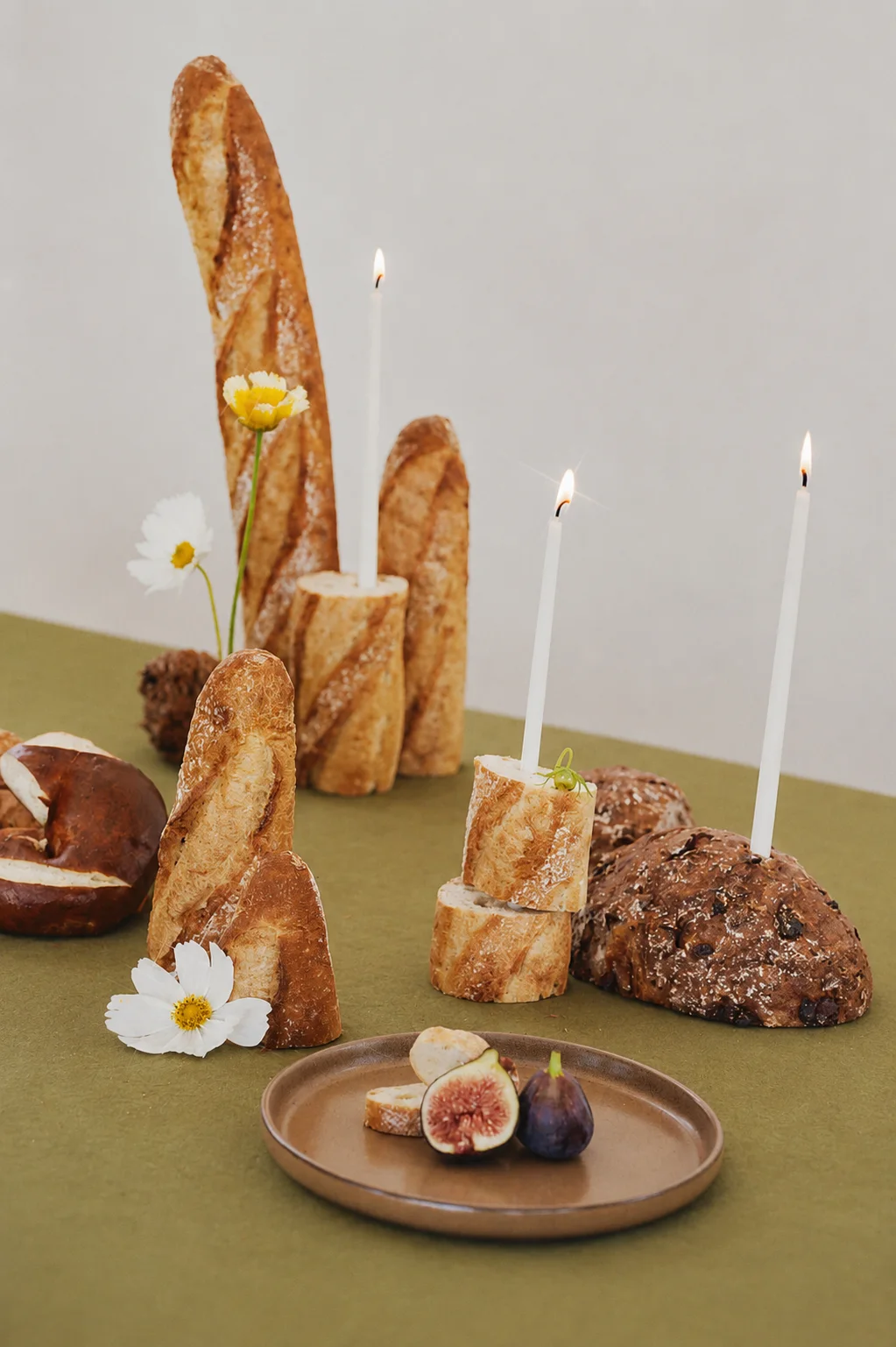 Upright baguettes and rustic bread with delicate white and yellow flowers, lit white taper candles, and a plate of sliced figs on a green tablecloth