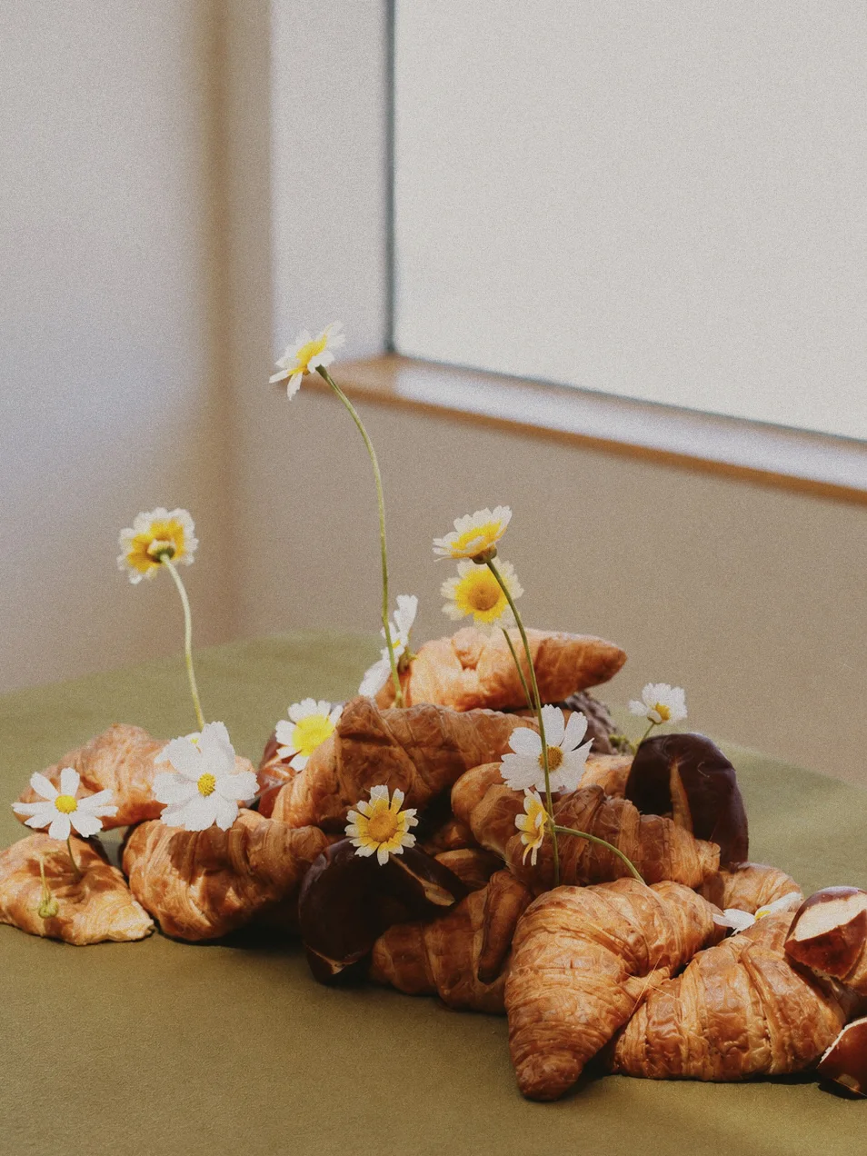 Side view of croissants and rustic loaves with white and yellow flowers on a green-covered table beside a window