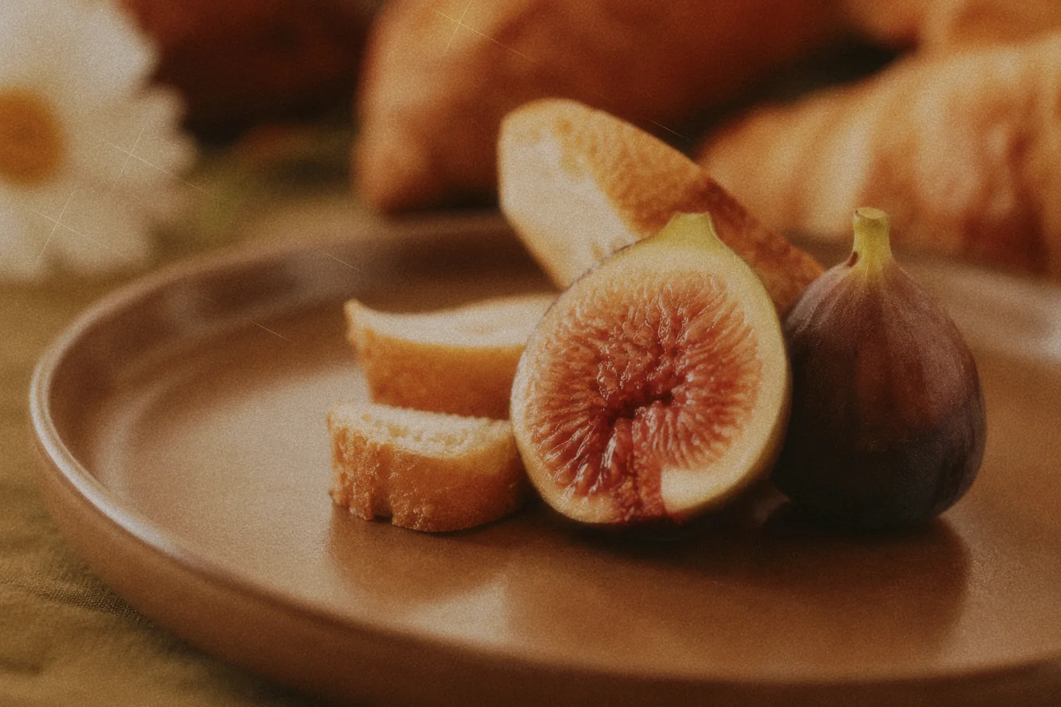 Halved fig and whole fig beside sliced bread on a brown ceramic plate