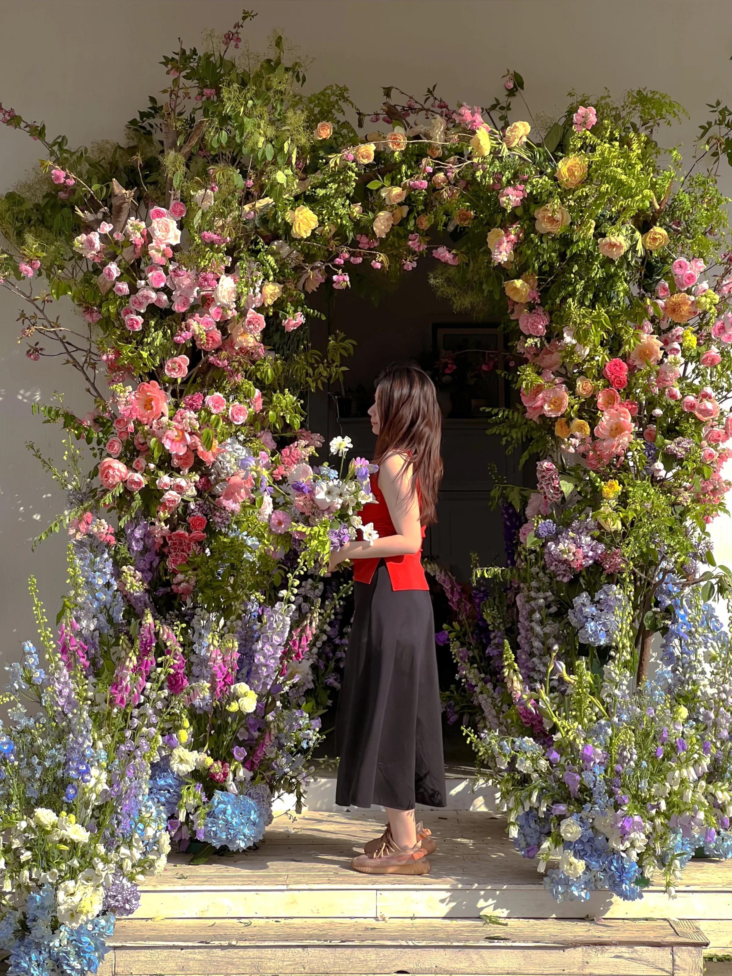 Florist standing within a full floral archway of roses, delphiniums, and hydrangeas framing a doorway