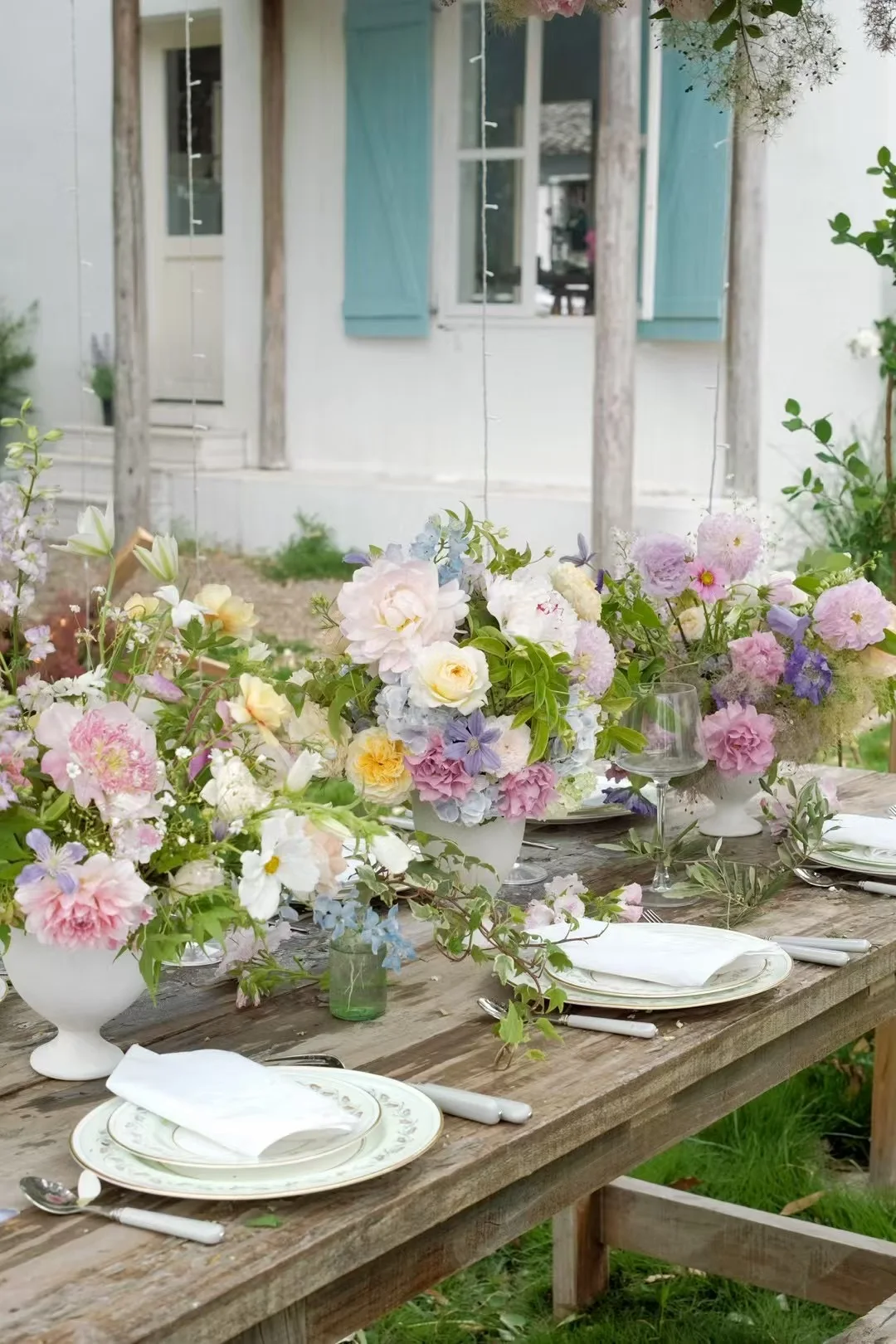 Rustic garden reception table lined with pastel centerpieces before a blue-shuttered cottage