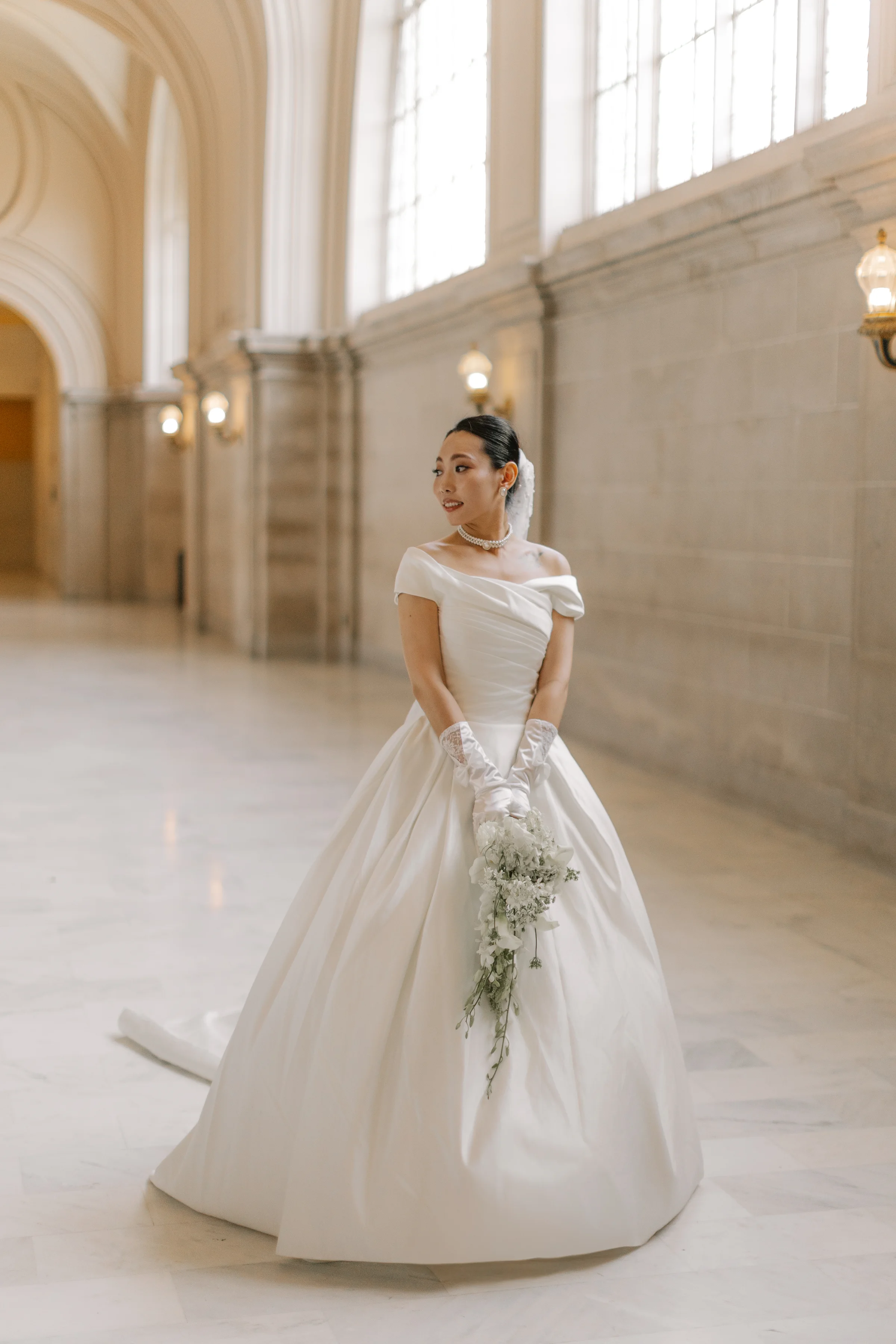 Bride in a sunlit marble hallway holding a cascading white bouquet