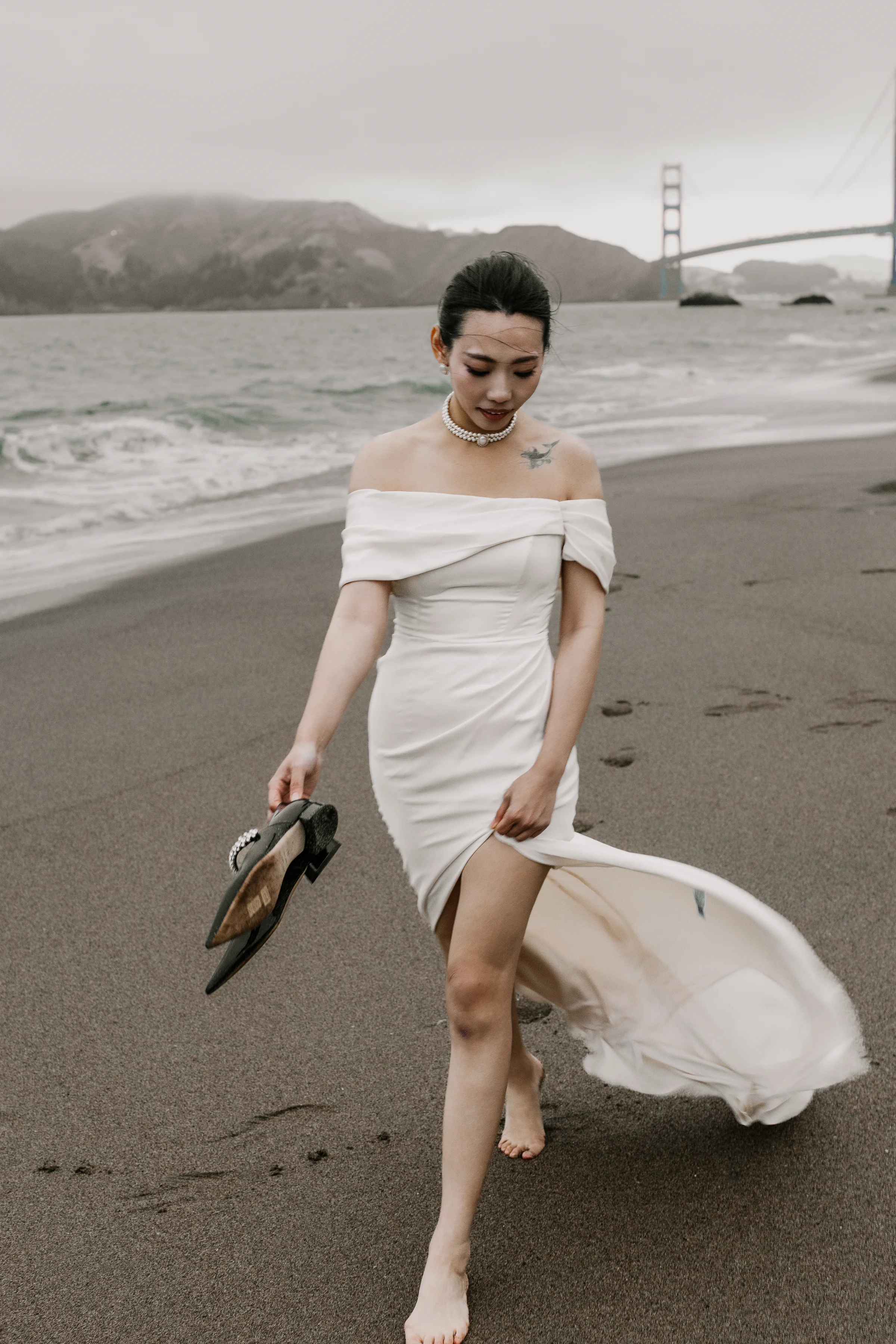 Bride in a short dress walking on the beach carrying her heels, the Golden Gate Bridge behind