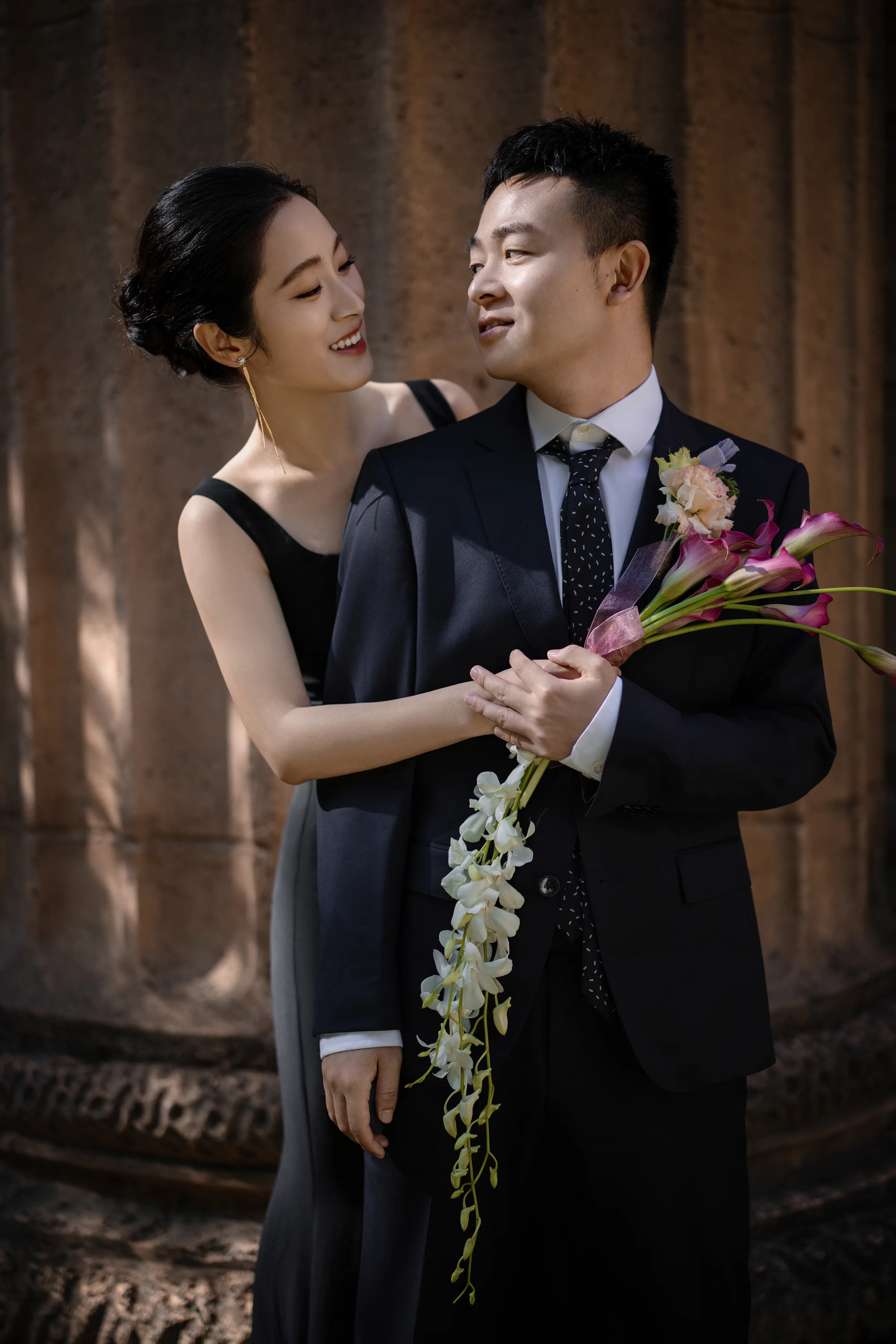 Bride in a black gown holding a pink orchid stem beside a classical column