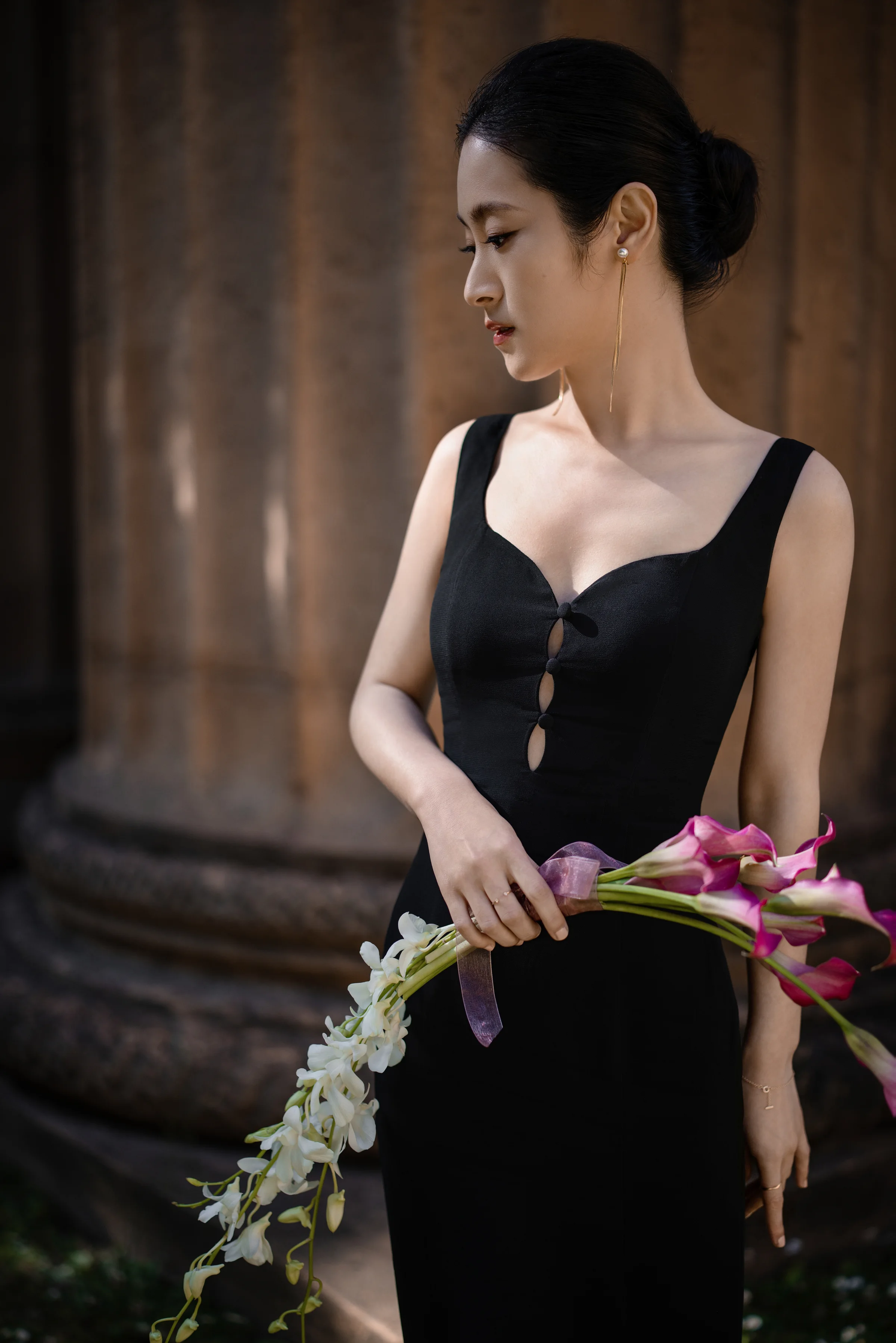 Bride in a black gown looking down while holding a pink orchid stem by a column