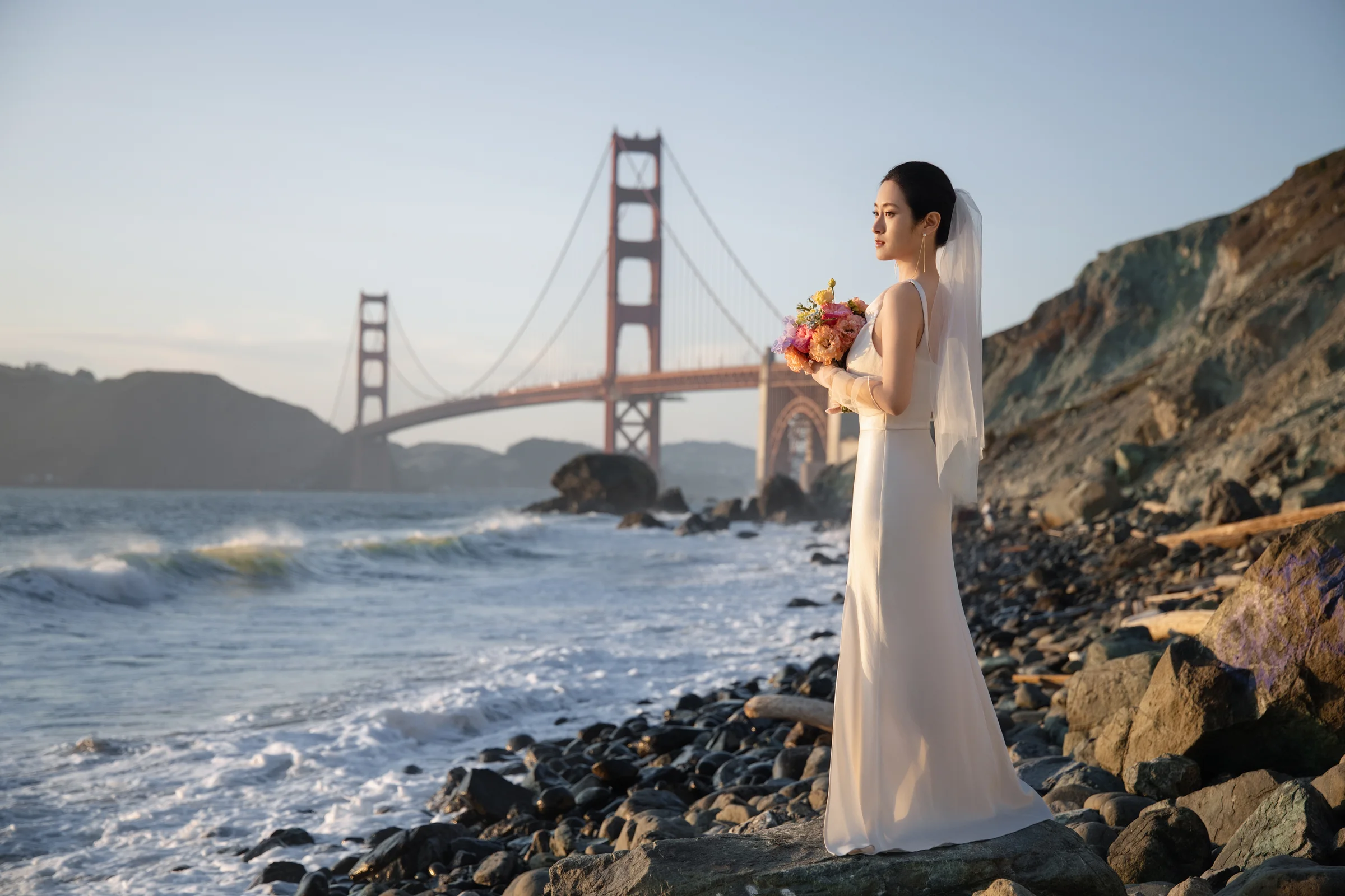 Bride in white holding a bouquet on the rocky shore with the Golden Gate Bridge behind