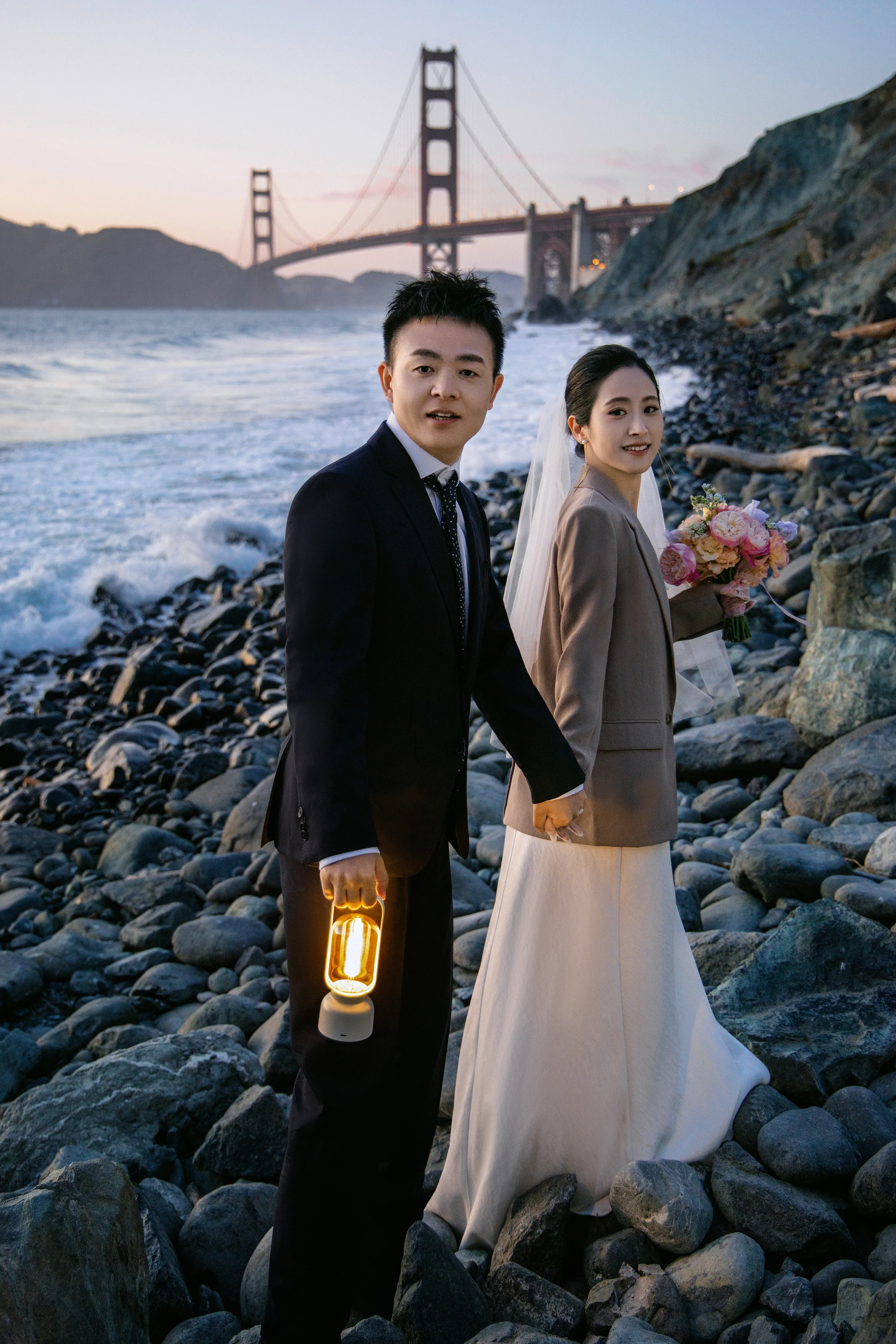 Groom holding a lantern beside the bride on the rocks at twilight, the bridge behind
