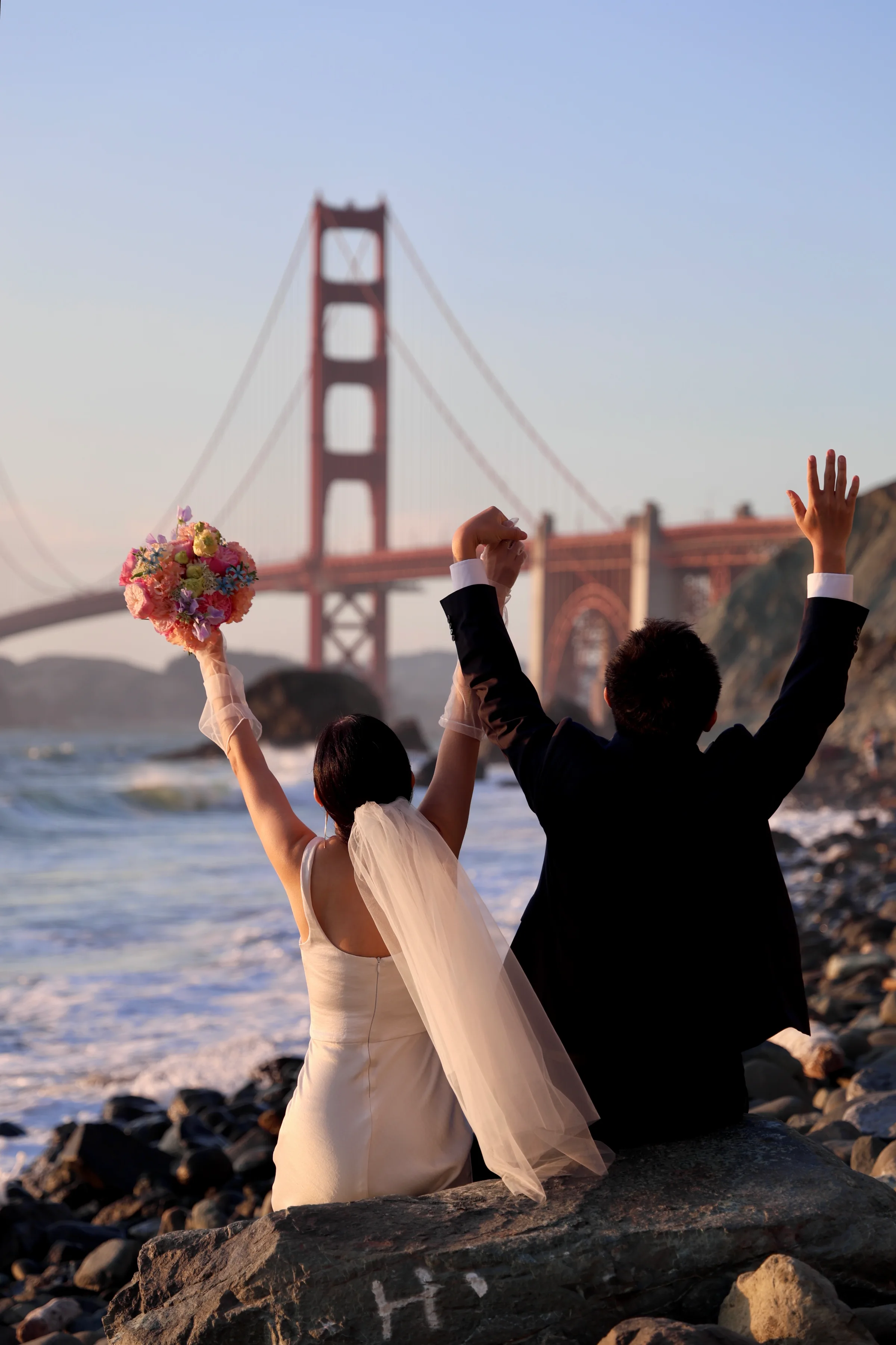 Couple seated on a rock with arms raised in celebration, the bridge behind