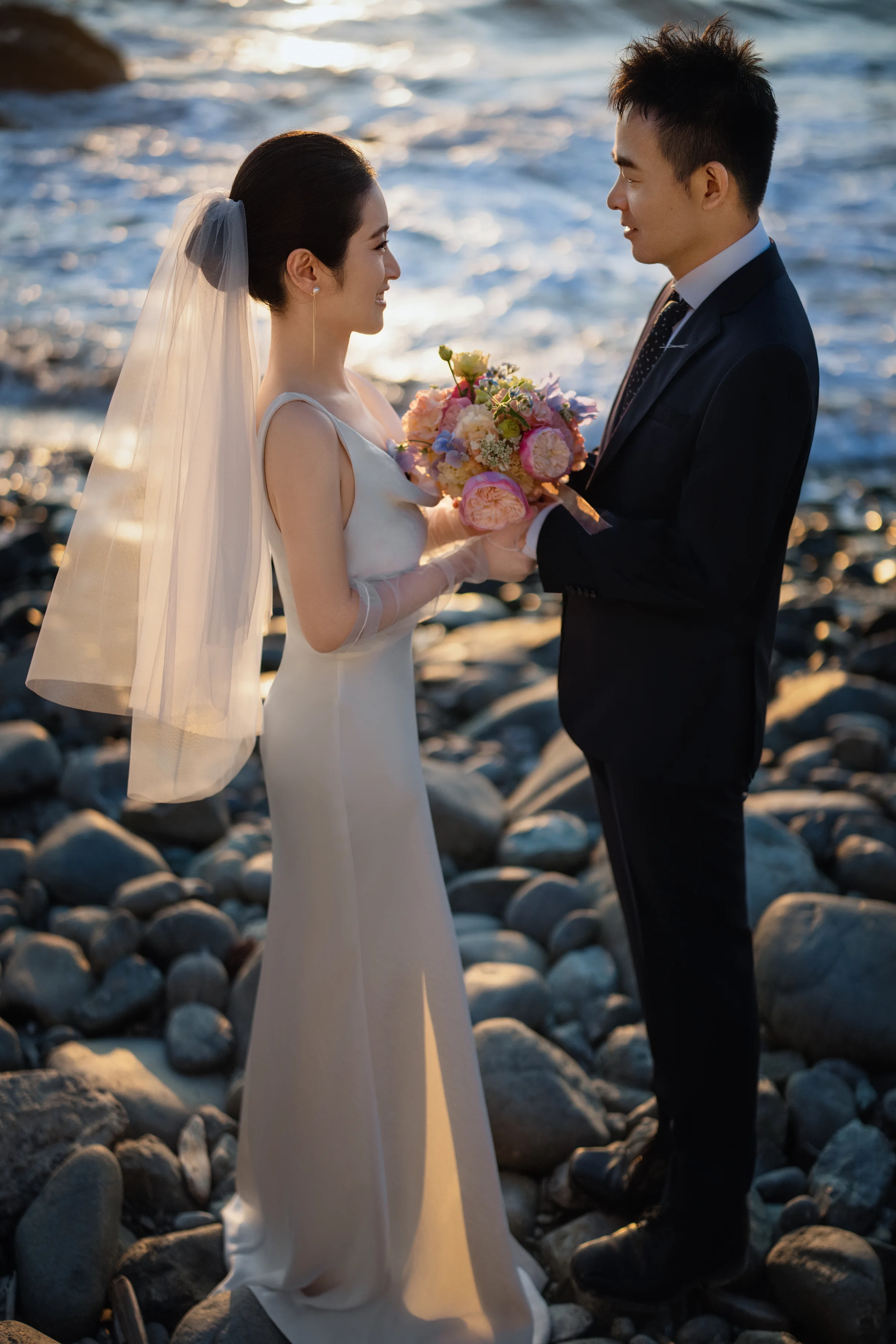 Couple facing each other on the beach, the bride holding a bouquet, the bridge behind