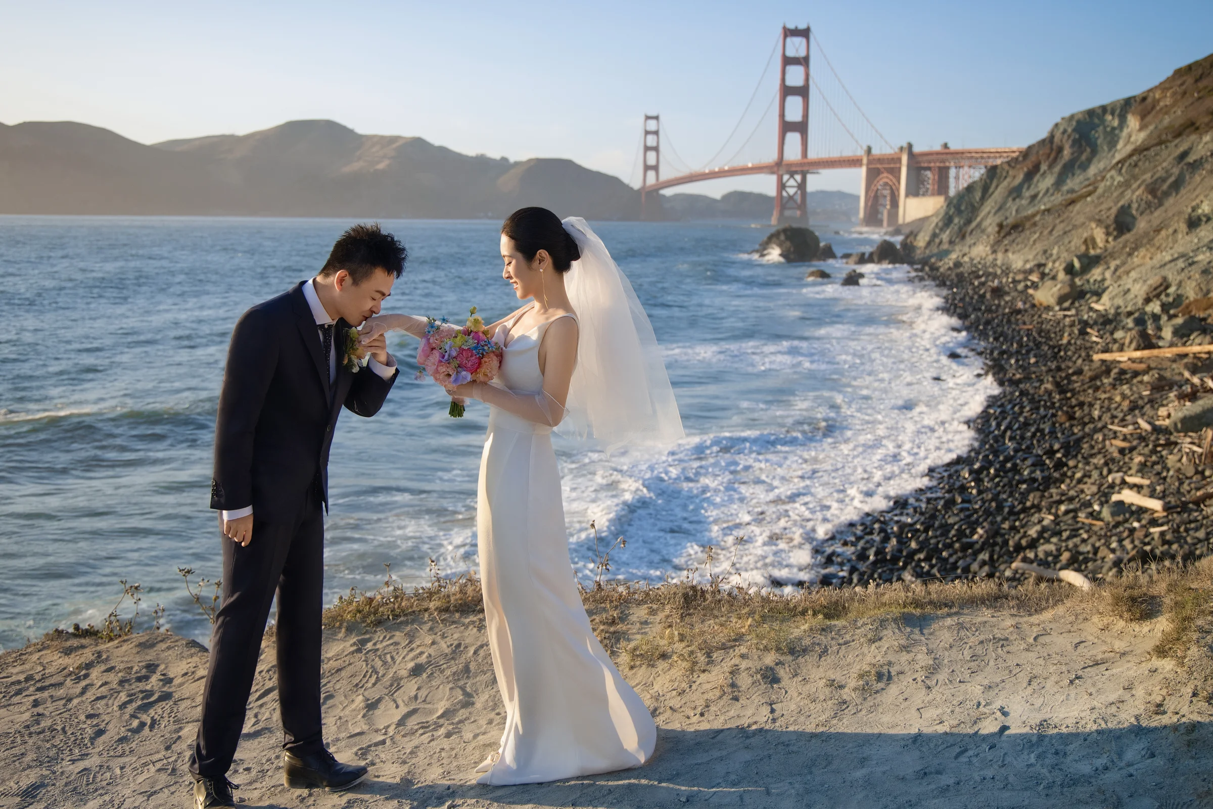 Groom kissing the bride's hand on the beach with the Golden Gate Bridge behind