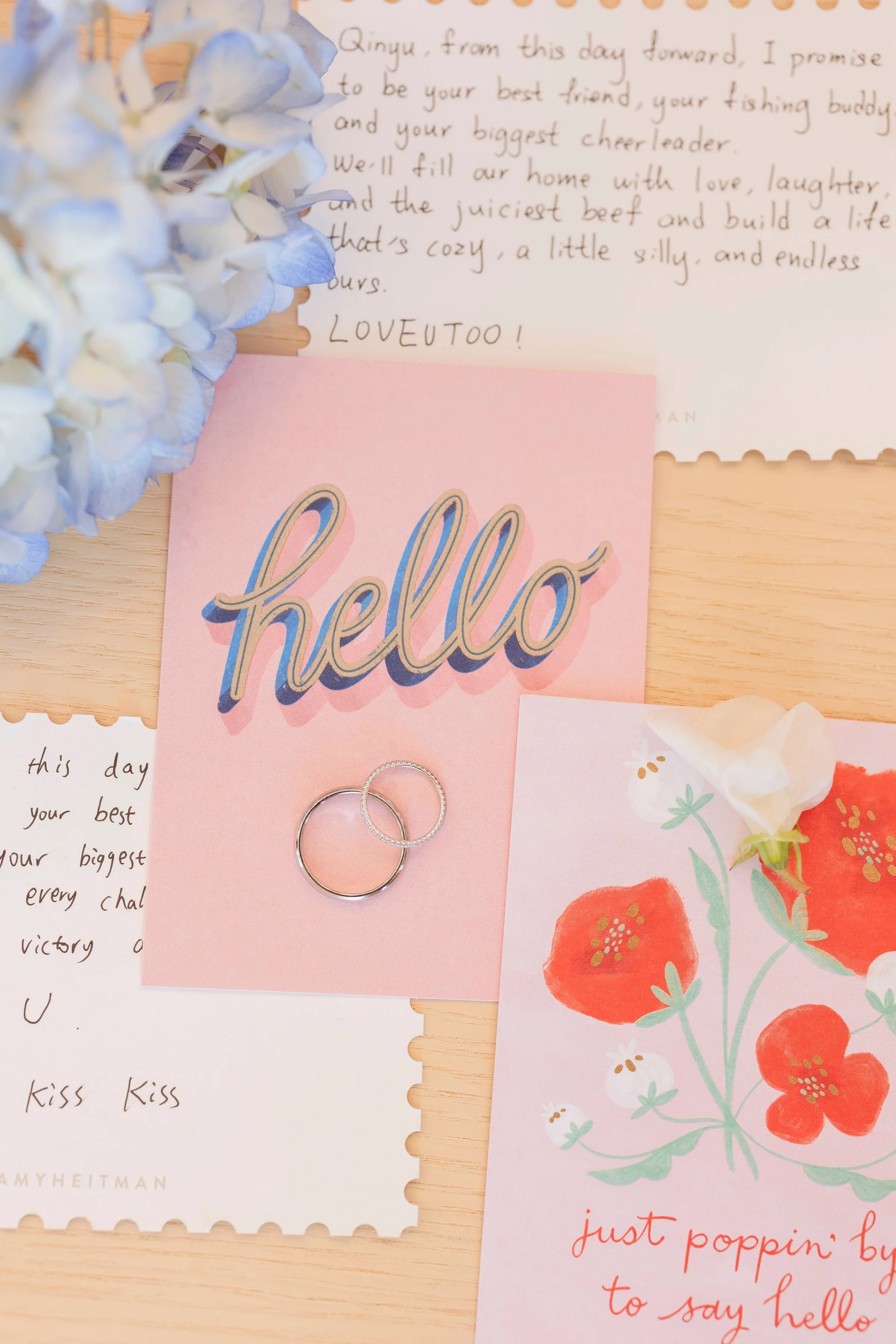 Flatlay of handwritten vow cards and two wedding rings beside blue hydrangea