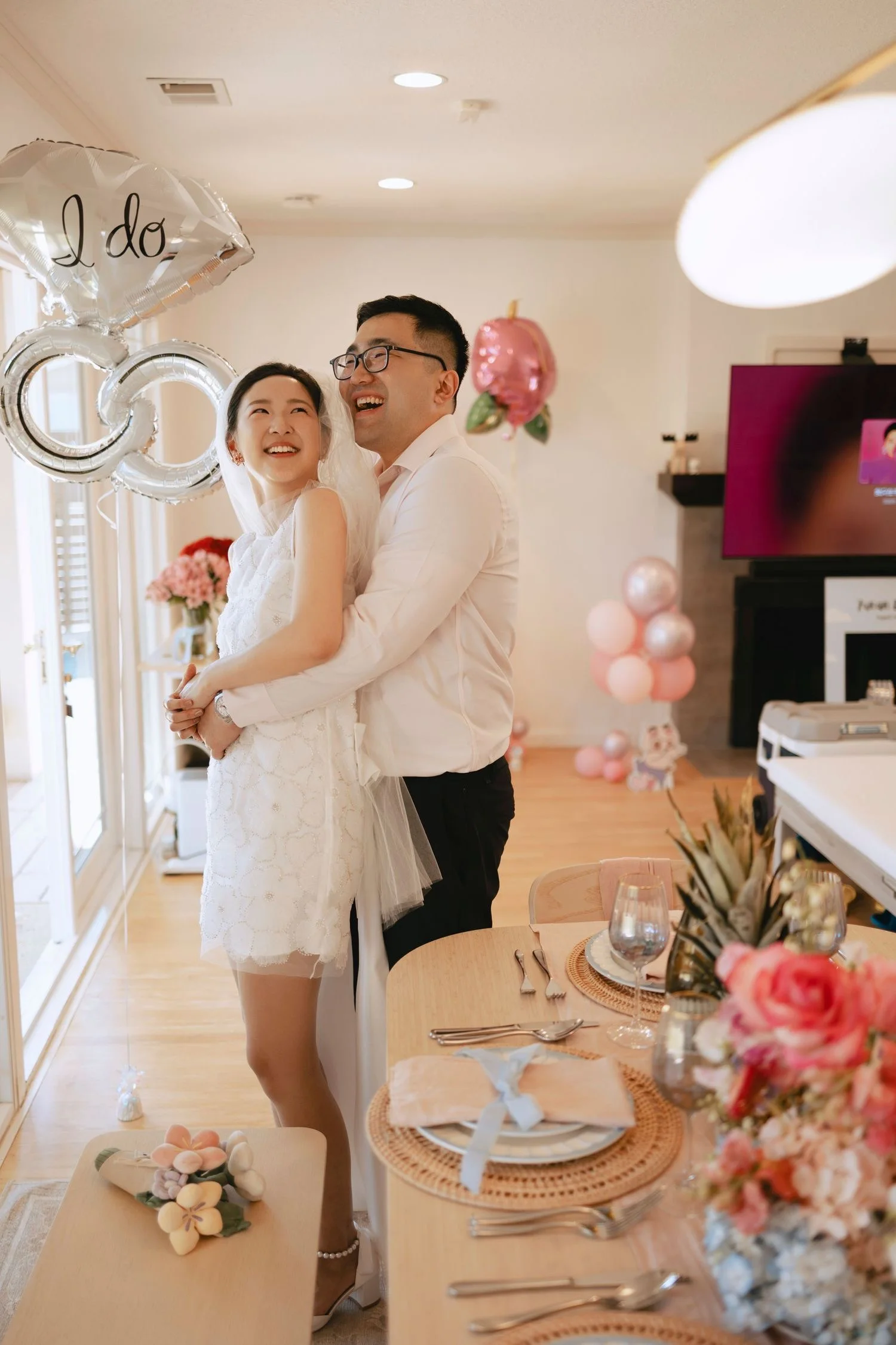 Couple embracing indoors beneath silver 'I do' ring balloons beside a styled dining table