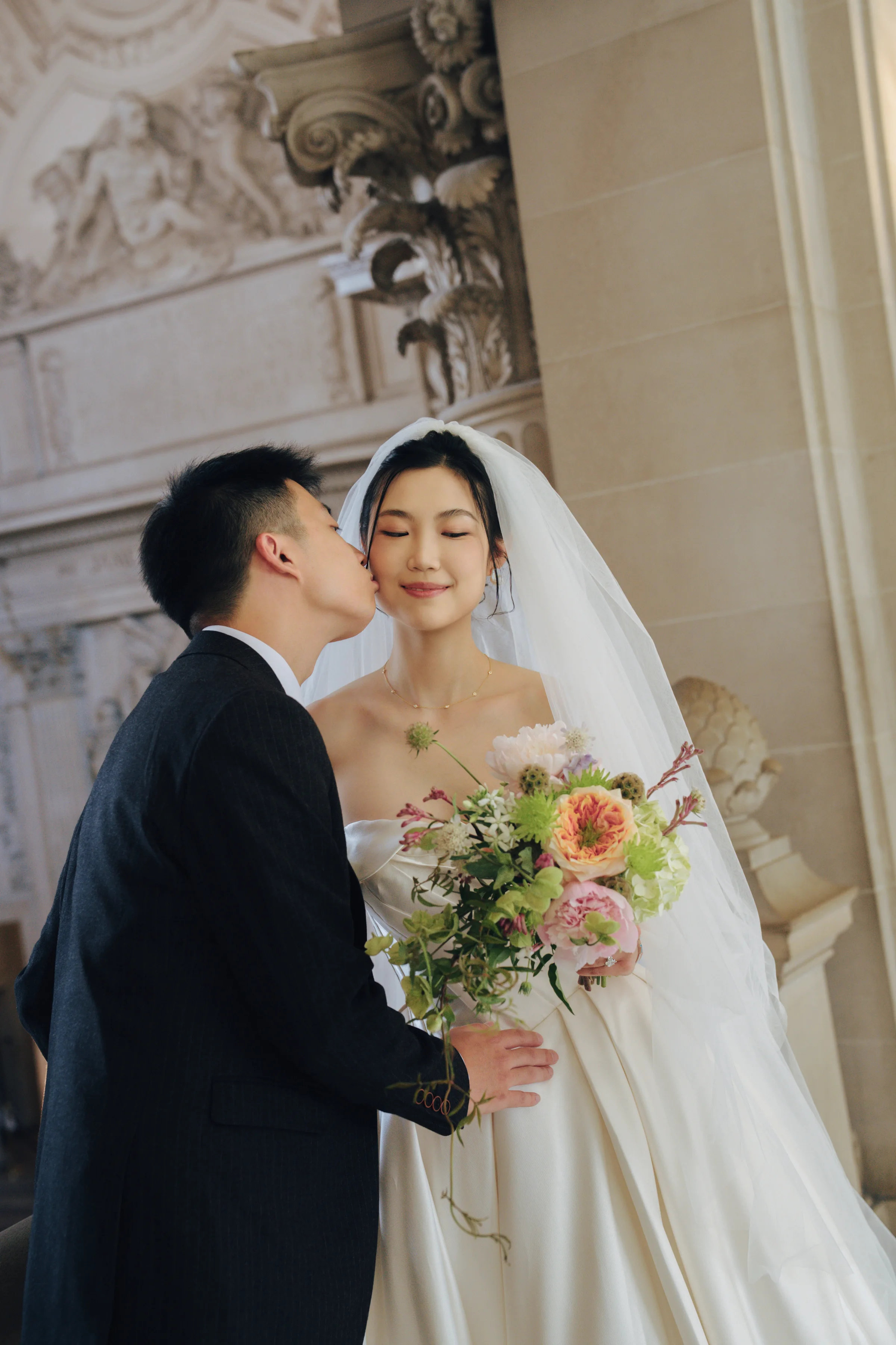 Groom kissing the bride's temple as she holds a peach and greenery bouquet