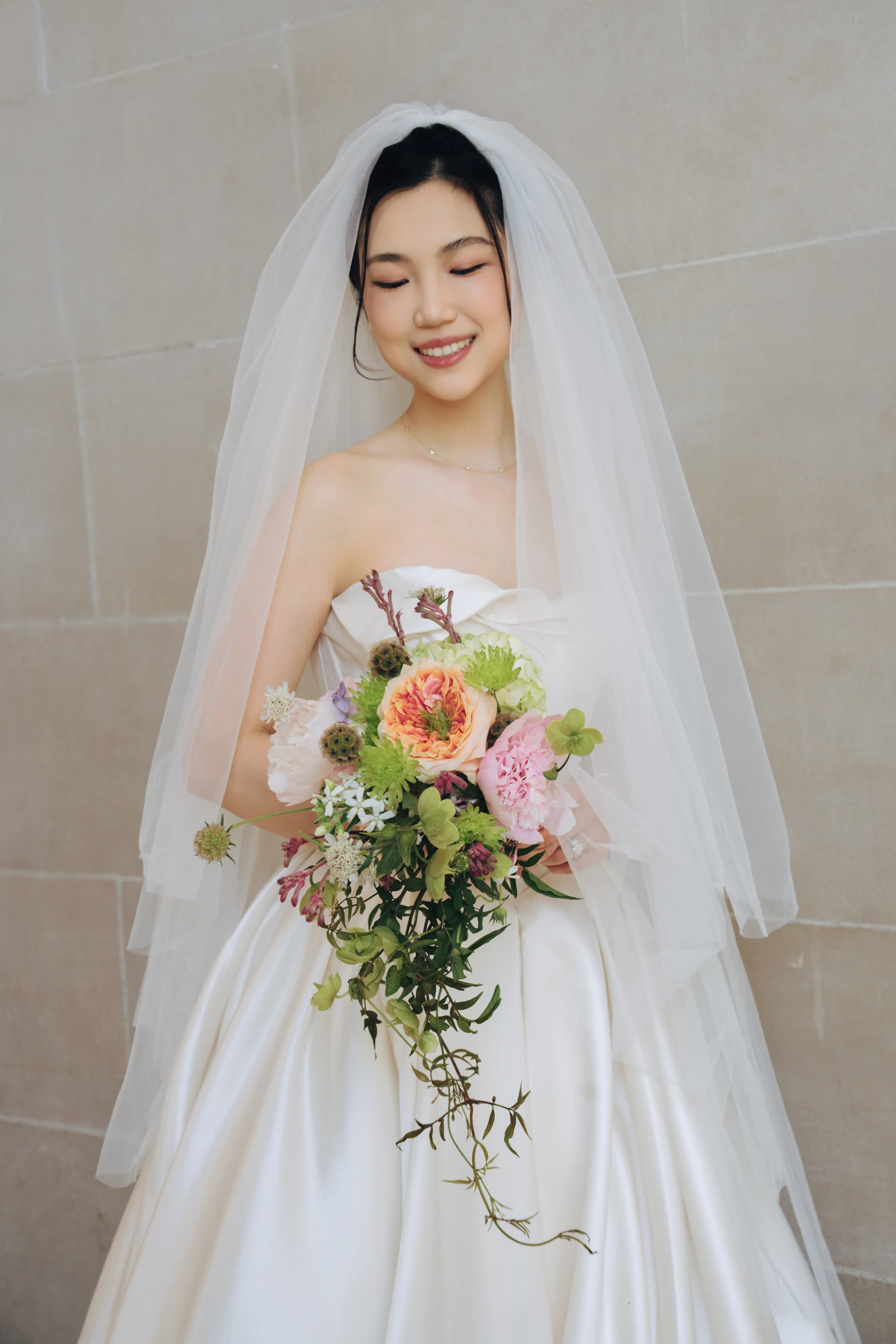 Bride against a marble wall holding a peach and greenery bouquet