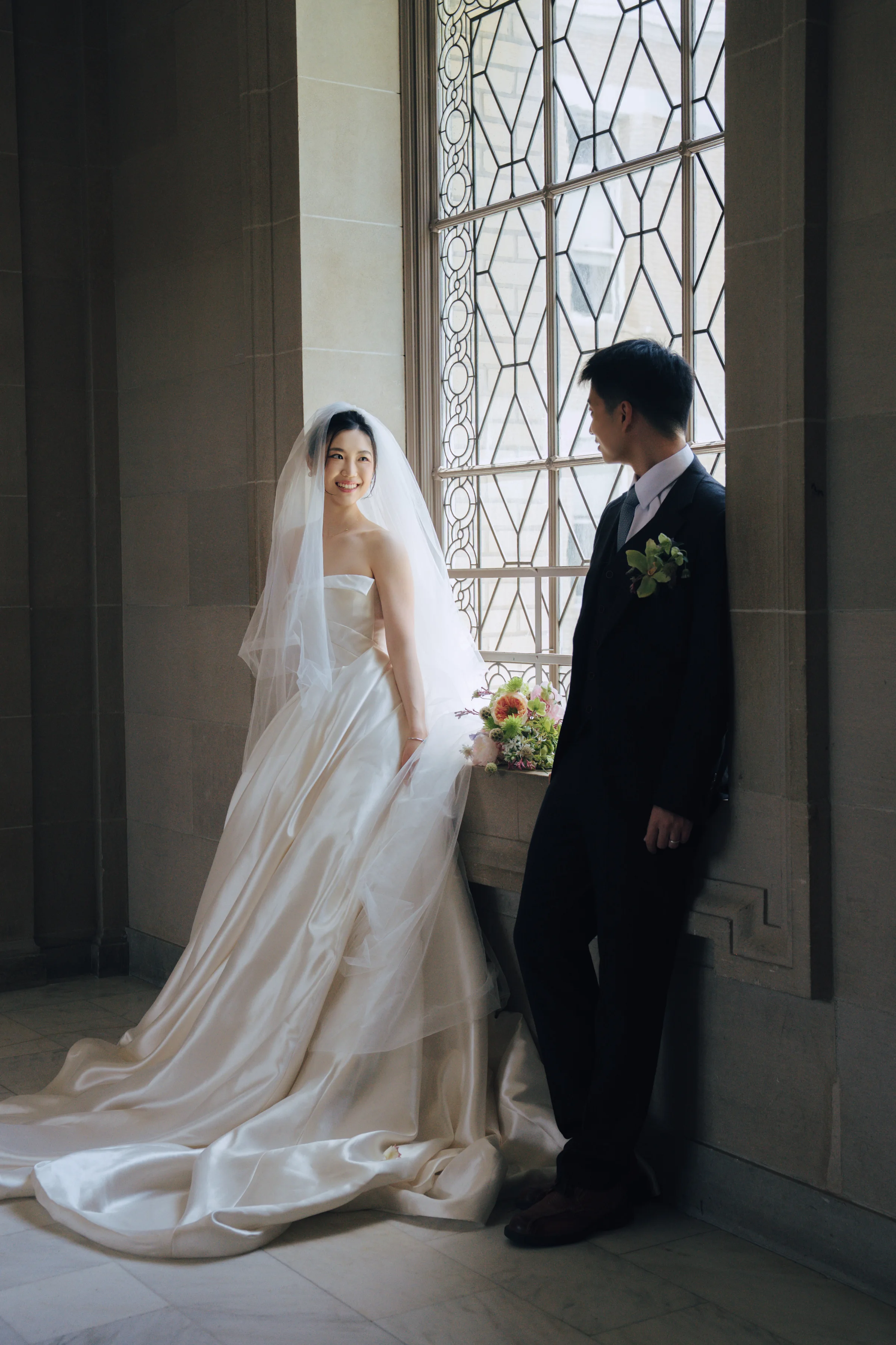Bride and groom by a bright lattice window, her train pooling on the floor