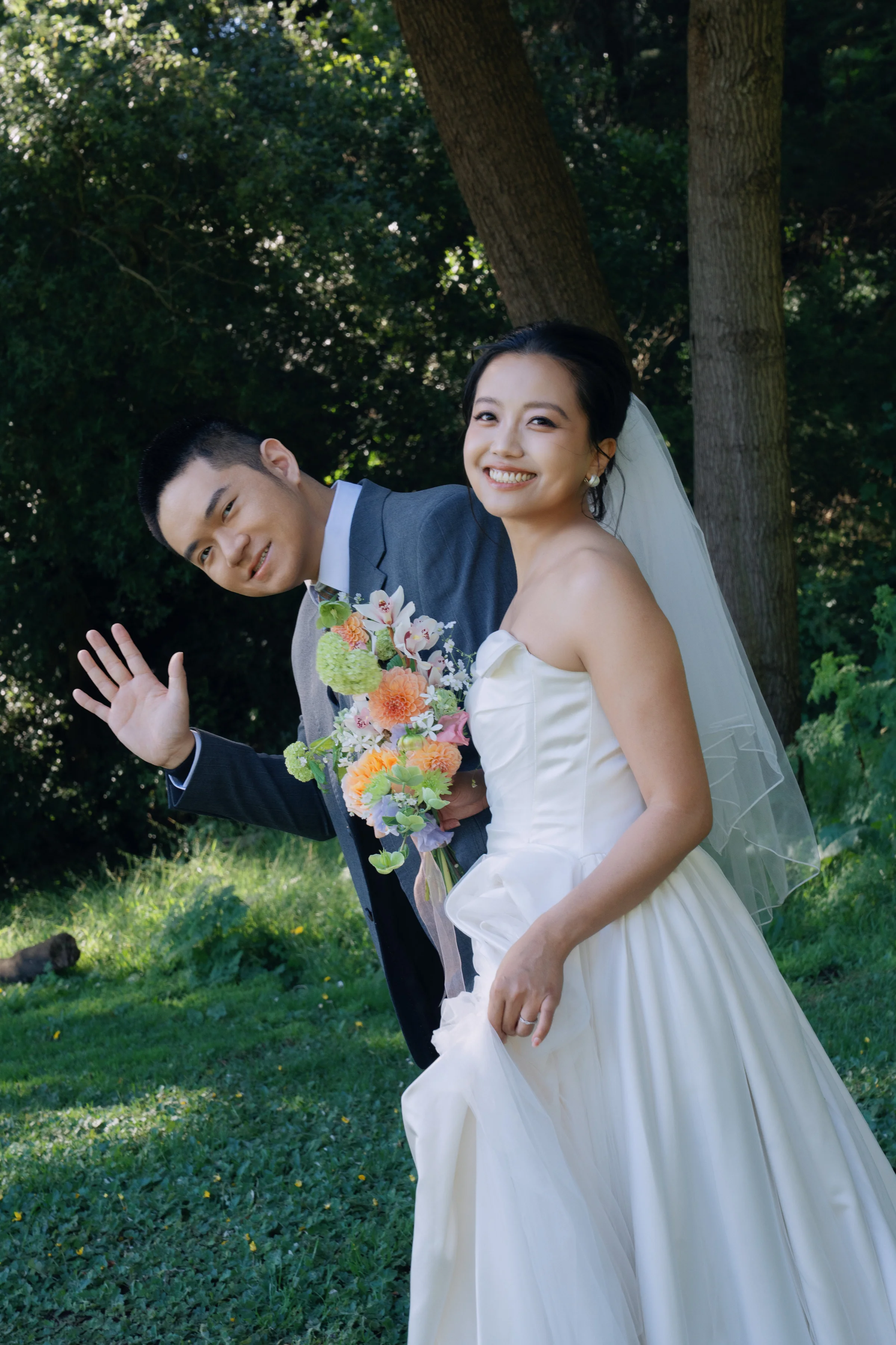 Groom waving playfully beside the smiling bride, who holds a peach bouquet under a tree