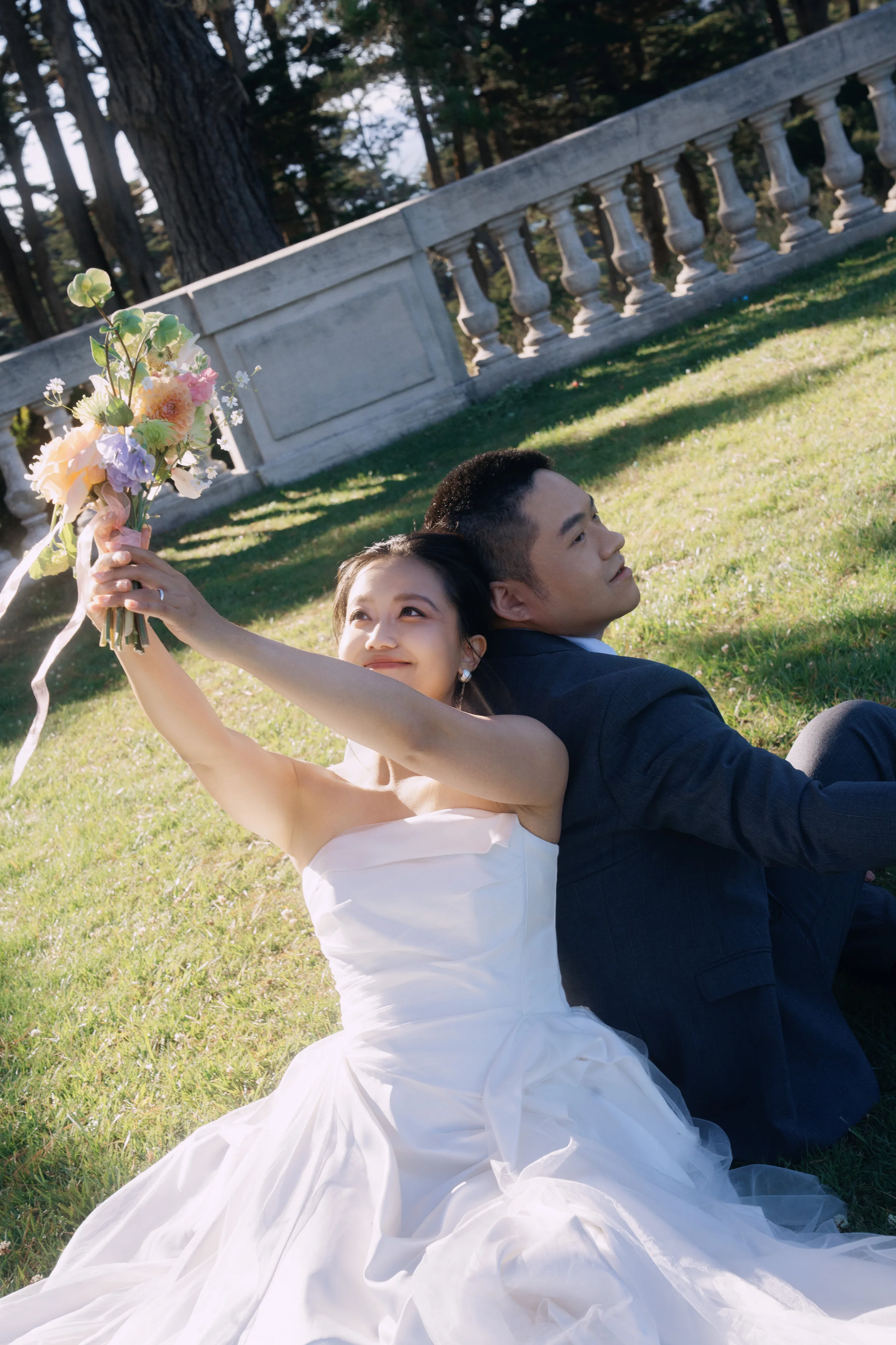 Bride holding her bouquet aloft as the groom reclines on the grass by a stone balustrade